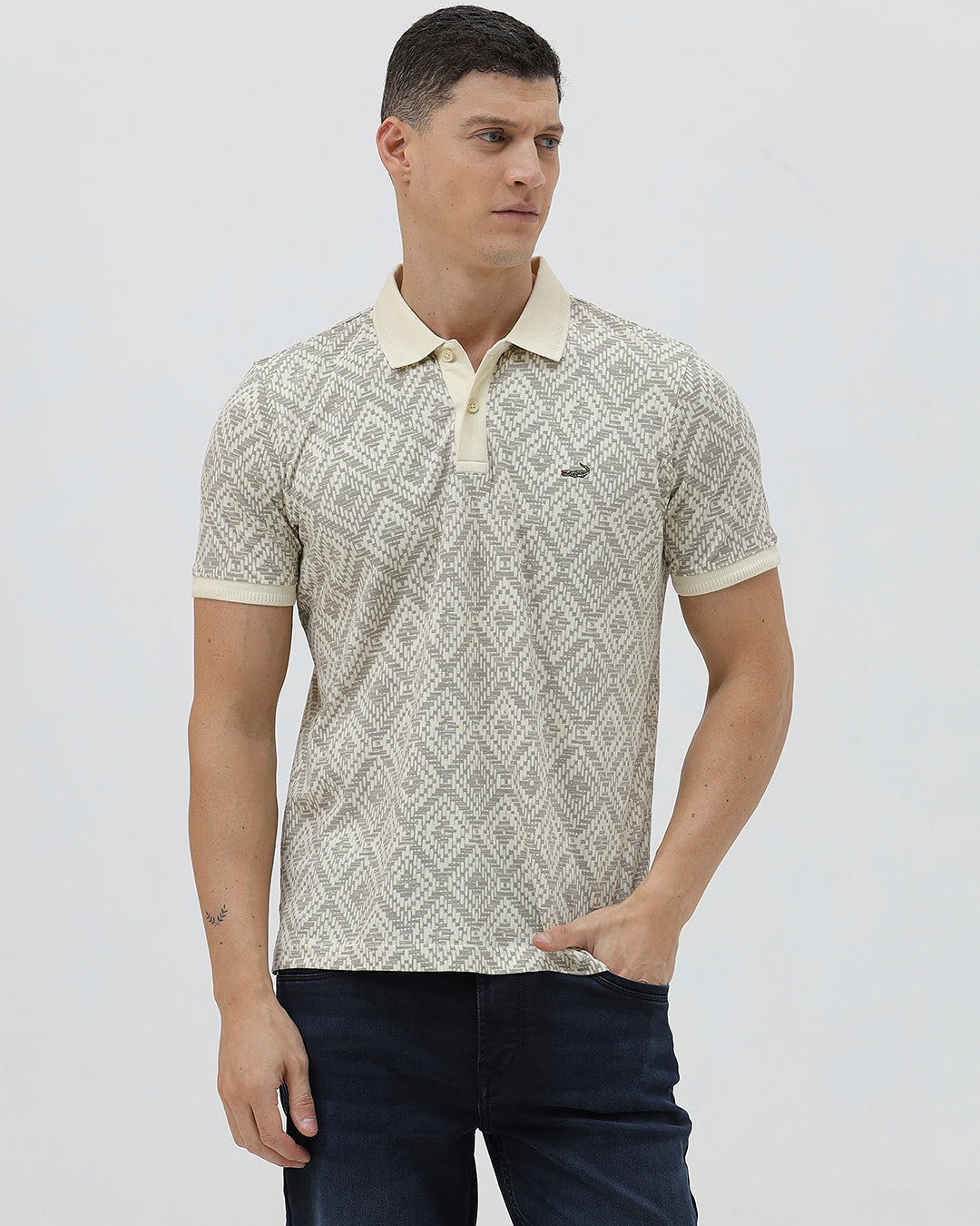 A man wearing a short-sleeve polo shirt with a beige and gray geometric pattern, paired with dark blue jeans, standing against a light gray background.
