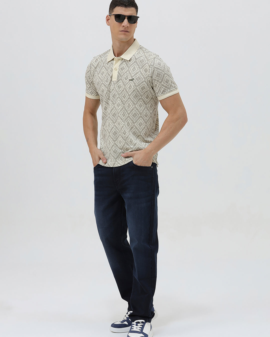 A man wearing a short-sleeve polo shirt with a beige and gray geometric pattern, paired with dark blue jeans, standing against a light gray background.