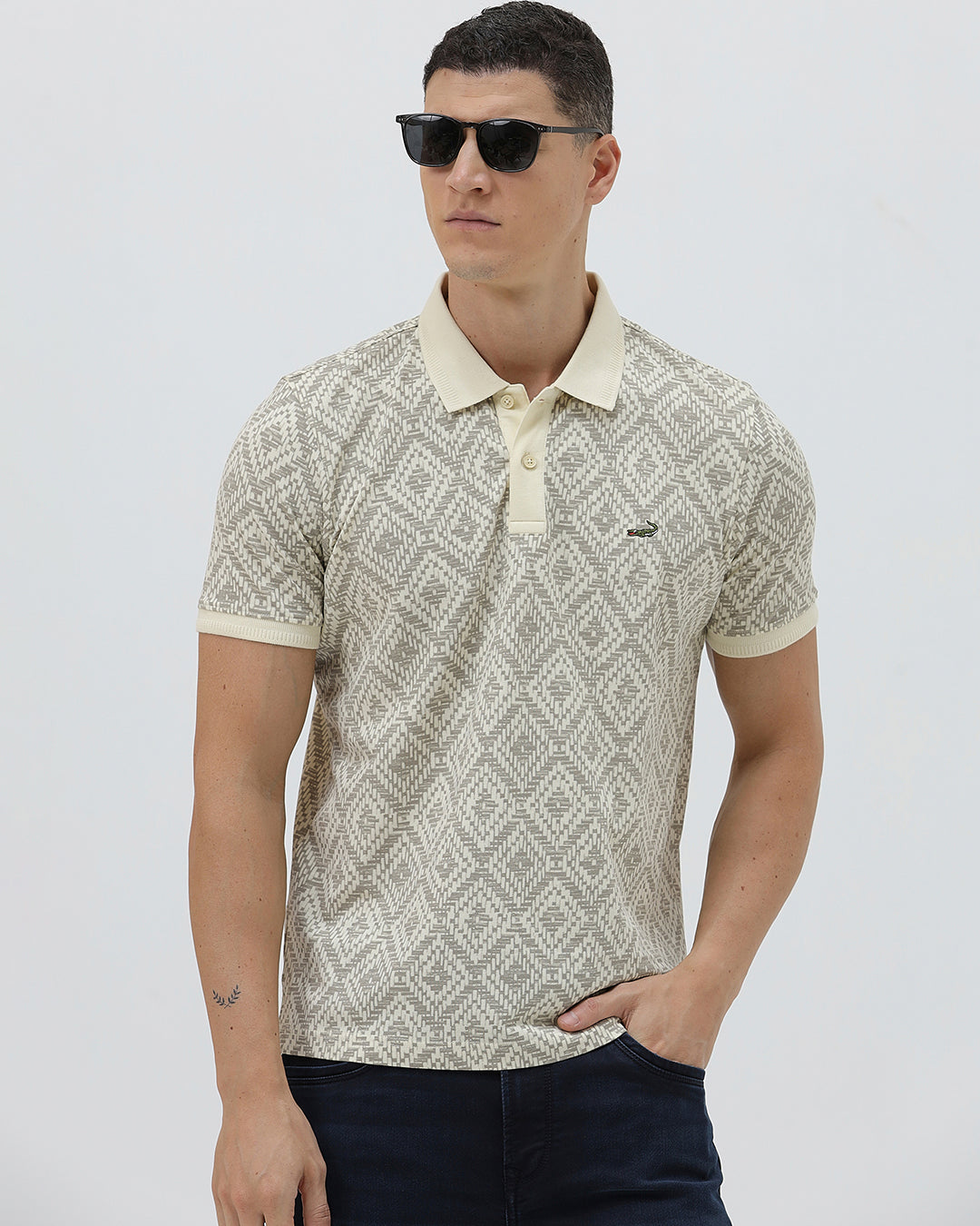 A man wearing a short-sleeve polo shirt with a beige and gray geometric pattern, paired with dark blue jeans, standing against a light gray background.
