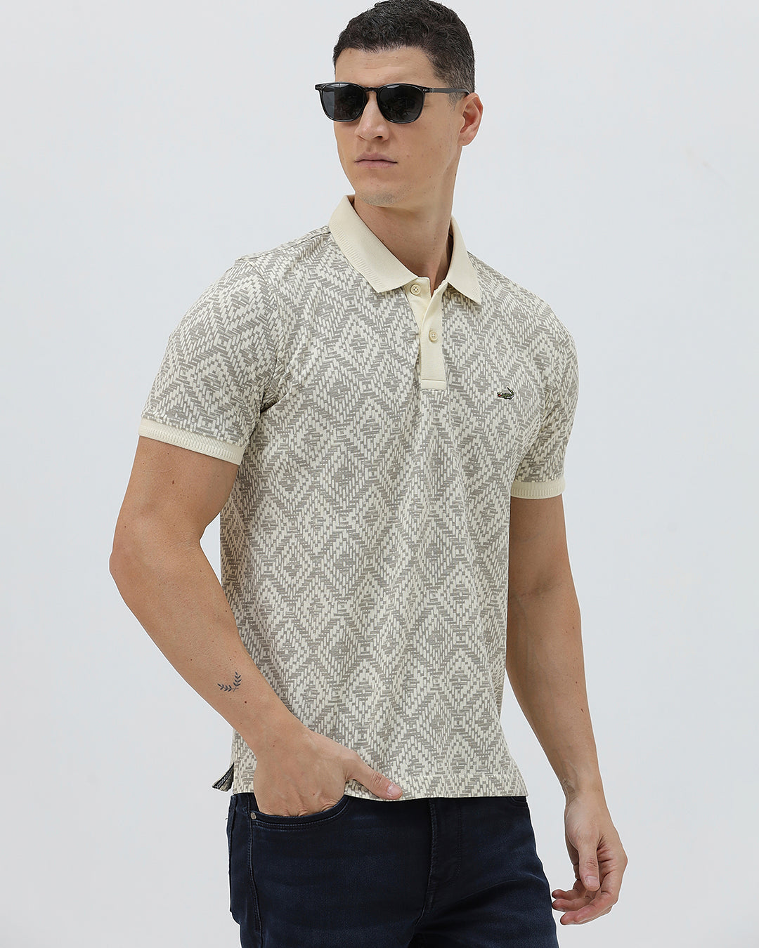 A man wearing a short-sleeve polo shirt with a beige and gray geometric pattern, paired with dark blue jeans, standing against a light gray background.