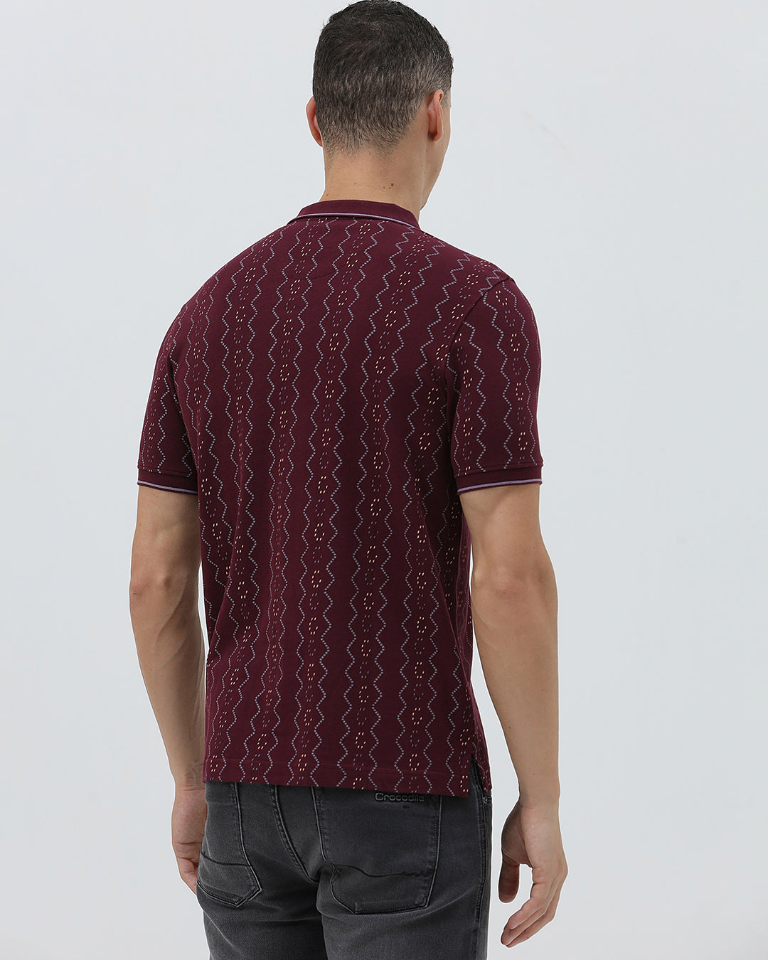 A man wearing a short-sleeve polo shirt in burgundy with a repeating vertical wavy pattern in dark and light red, paired with dark gray jeans, against a plain light gray background.