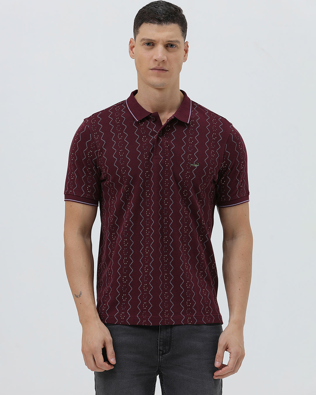 A man wearing a short-sleeve polo shirt in burgundy with a repeating vertical wavy pattern in dark and light red, paired with dark gray jeans, against a plain light gray background.