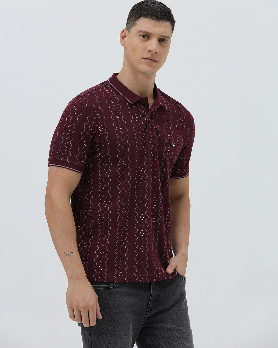 A man wearing a short-sleeve polo shirt in burgundy with a repeating vertical wavy pattern in dark and light red, paired with dark gray jeans, against a plain light gray background.