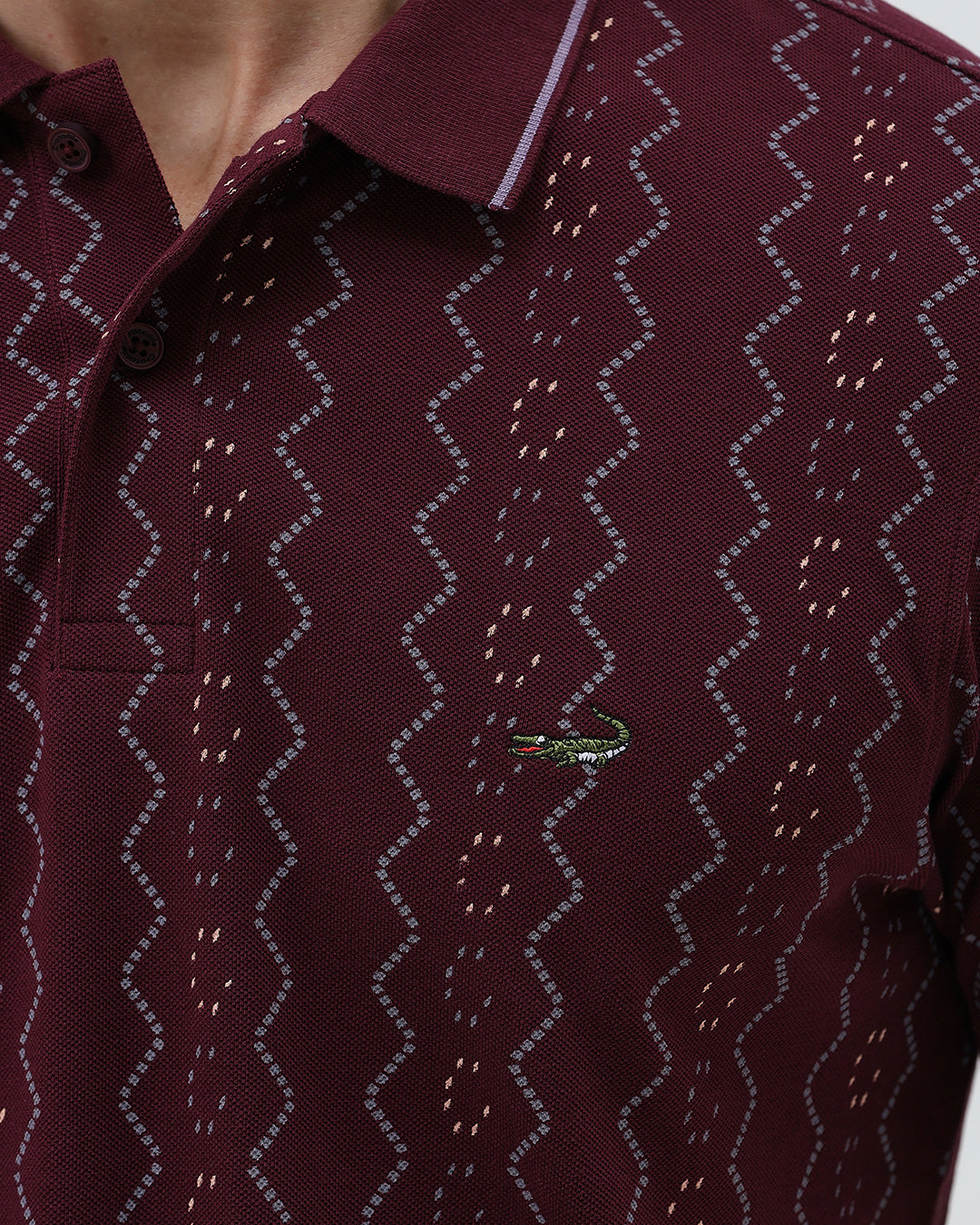 A man wearing a short-sleeve polo shirt in burgundy with a repeating vertical wavy pattern in dark and light red, paired with dark gray jeans, against a plain light gray background.