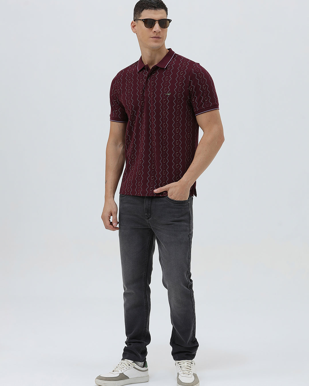 A man wearing a short-sleeve polo shirt in burgundy with a repeating vertical wavy pattern in dark and light red, paired with dark gray jeans, against a plain light gray background.