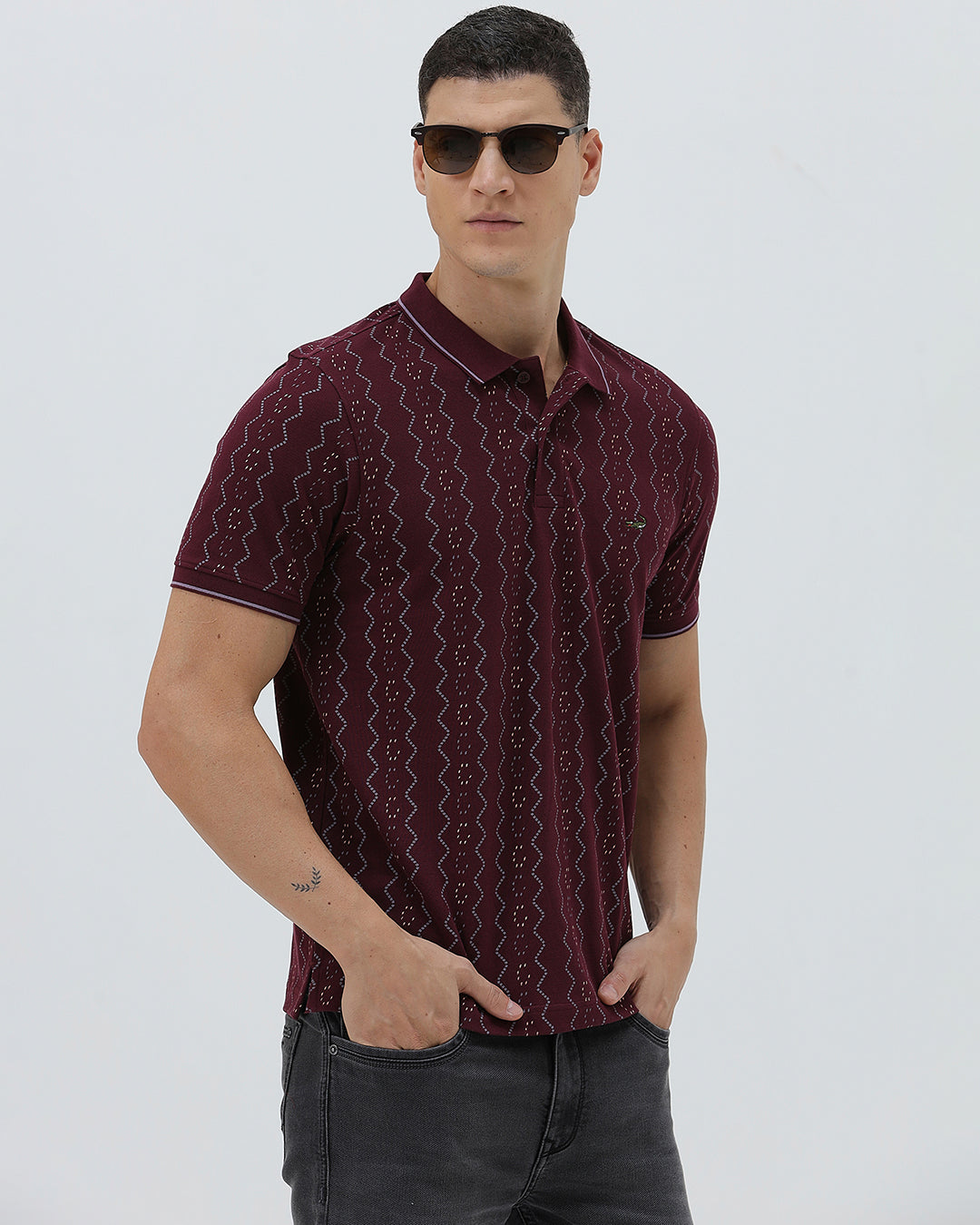 A man wearing a short-sleeve polo shirt in burgundy with a repeating vertical wavy pattern in dark and light red, paired with dark gray jeans, against a plain light gray background.