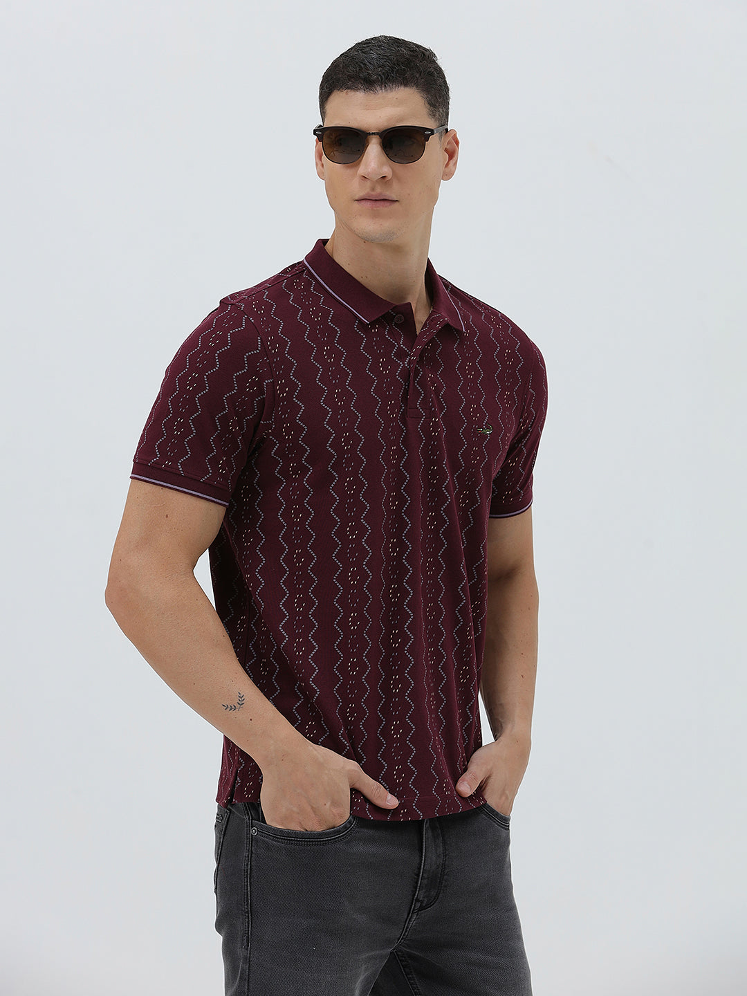 A man wearing a short-sleeve polo shirt in burgundy with a repeating vertical wavy pattern in dark and light red, paired with dark gray jeans, against a plain light gray background.