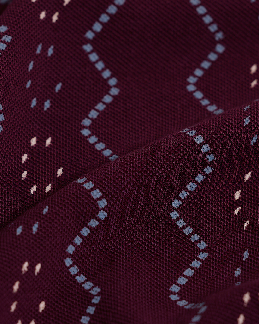 A man wearing a short-sleeve polo shirt in burgundy with a repeating vertical wavy pattern in dark and light red, paired with dark gray jeans, against a plain light gray background.