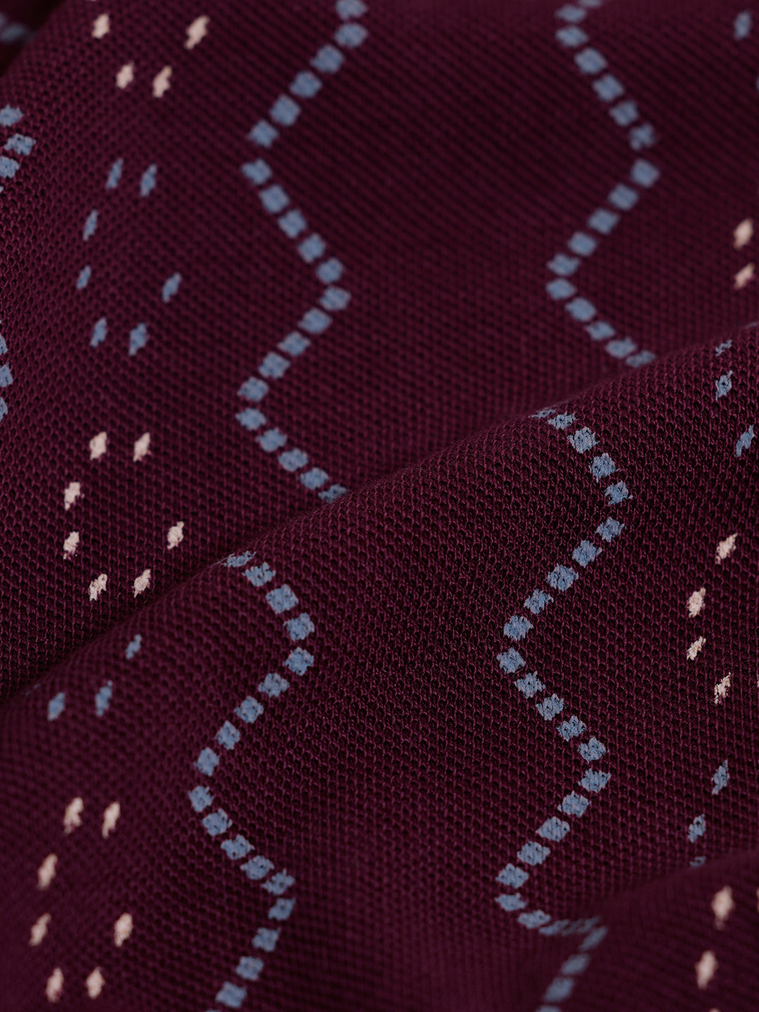 A man wearing a short-sleeve polo shirt in burgundy with a repeating vertical wavy pattern in dark and light red, paired with dark gray jeans, against a plain light gray background.