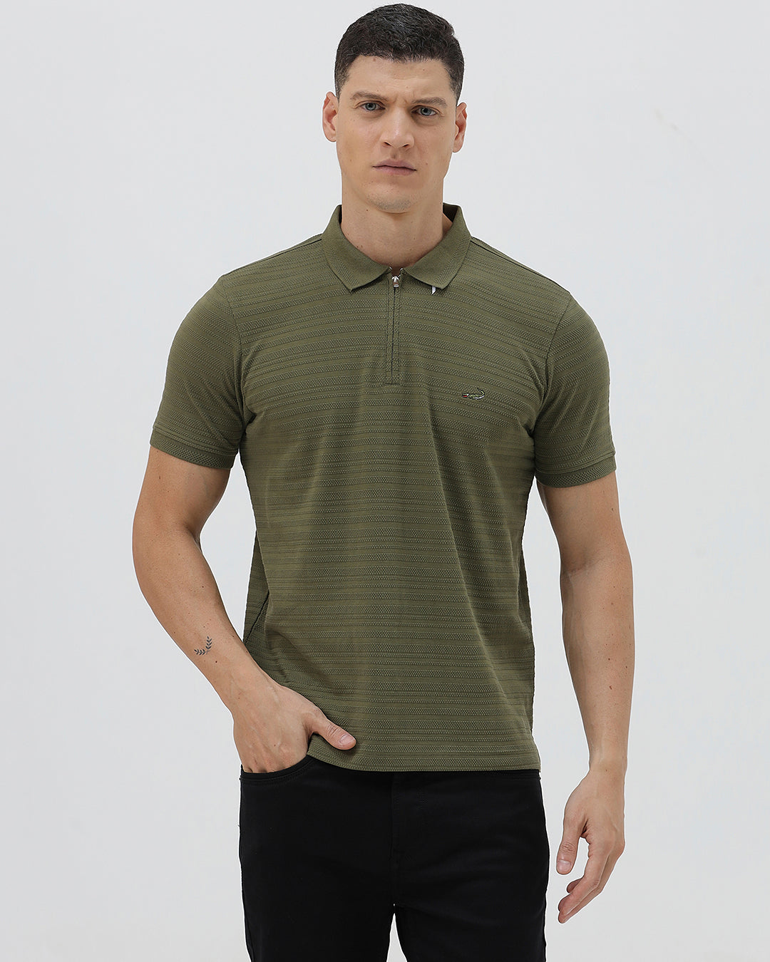 A man wearing an olive green short-sleeve polo shirt with a half-zip collar and black pants, standing against a light gray studio background.