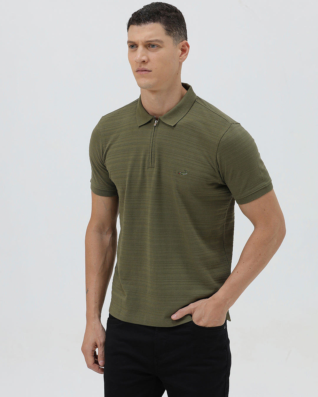 A man wearing an olive green short-sleeve polo shirt with a half-zip collar and black pants, standing against a light gray studio background.