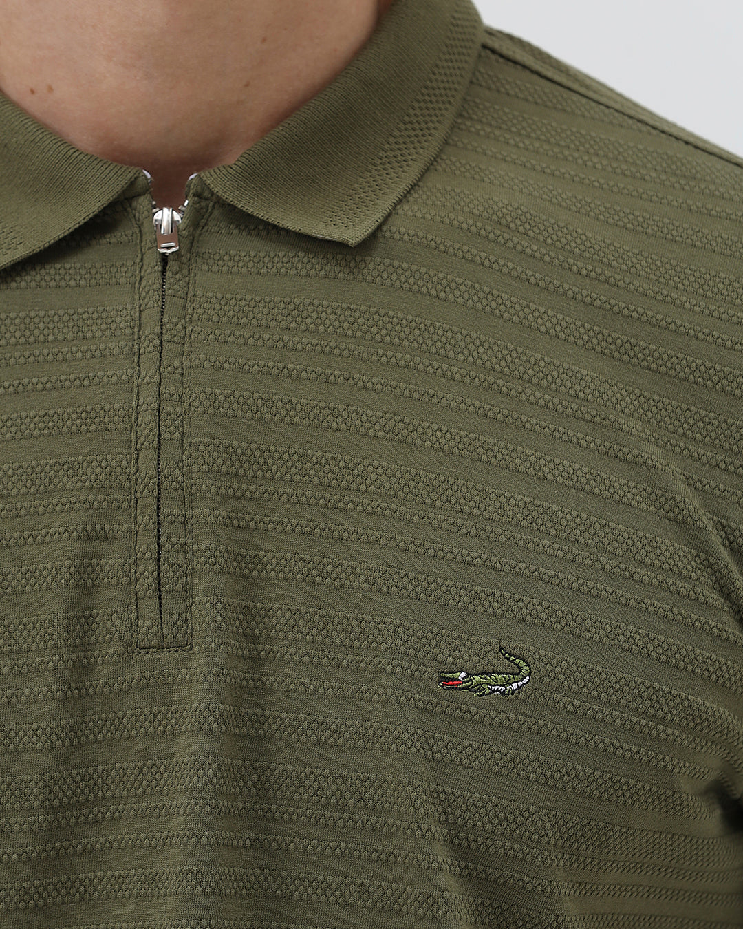 A man wearing an olive green short-sleeve polo shirt with a half-zip collar and black pants, standing against a light gray studio background.