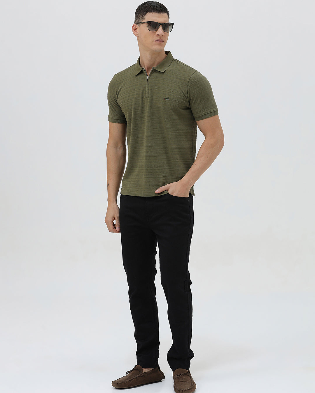 A man wearing an olive green short-sleeve polo shirt with a half-zip collar and black pants, standing against a light gray studio background.