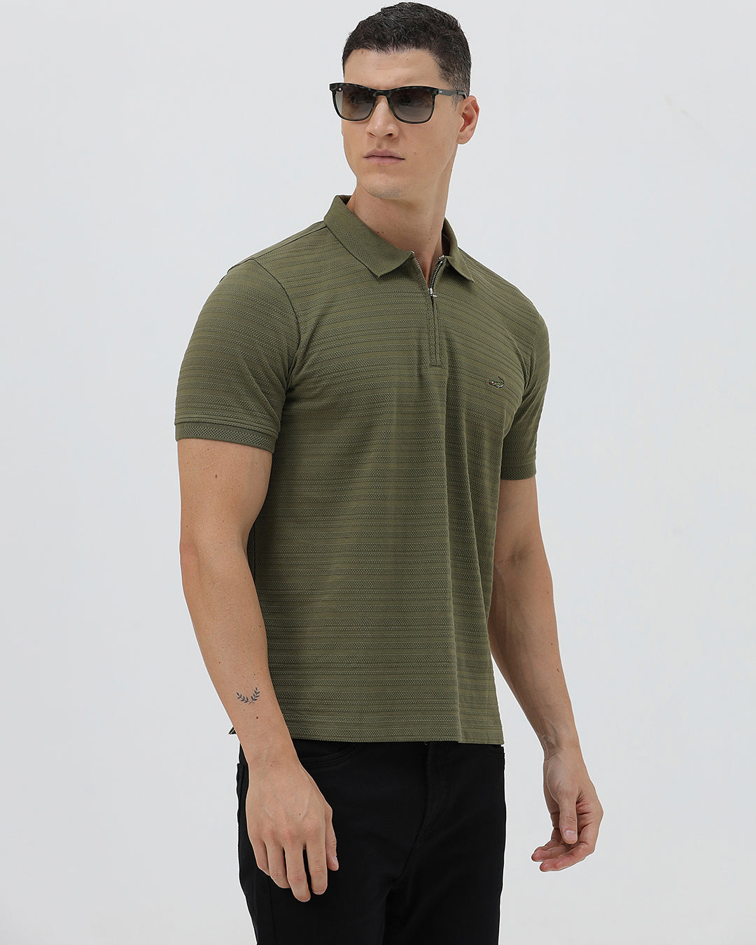 A man wearing an olive green short-sleeve polo shirt with a half-zip collar and black pants, standing against a light gray studio background.