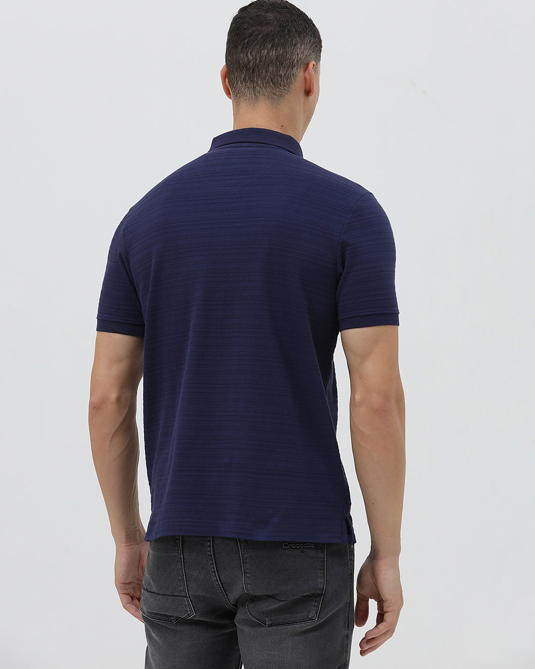 A man wearing a navy blue short-sleeve polo shirt with a small embroidered crocodile logo on the chest, paired with dark gray jeans, against a light gray studio background.