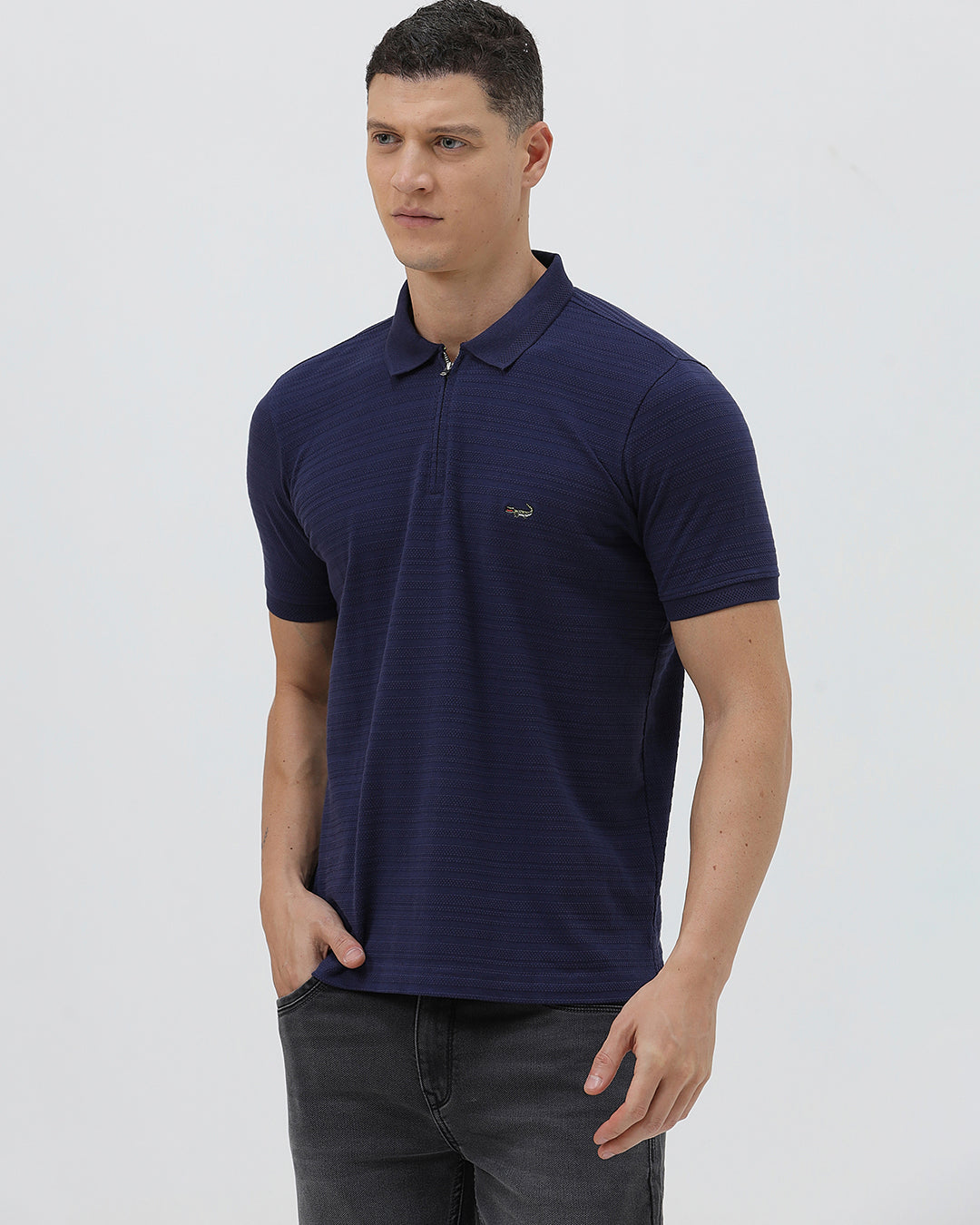 A man wearing a navy blue short-sleeve polo shirt with a small embroidered crocodile logo on the chest, paired with dark gray jeans, against a light gray studio background.