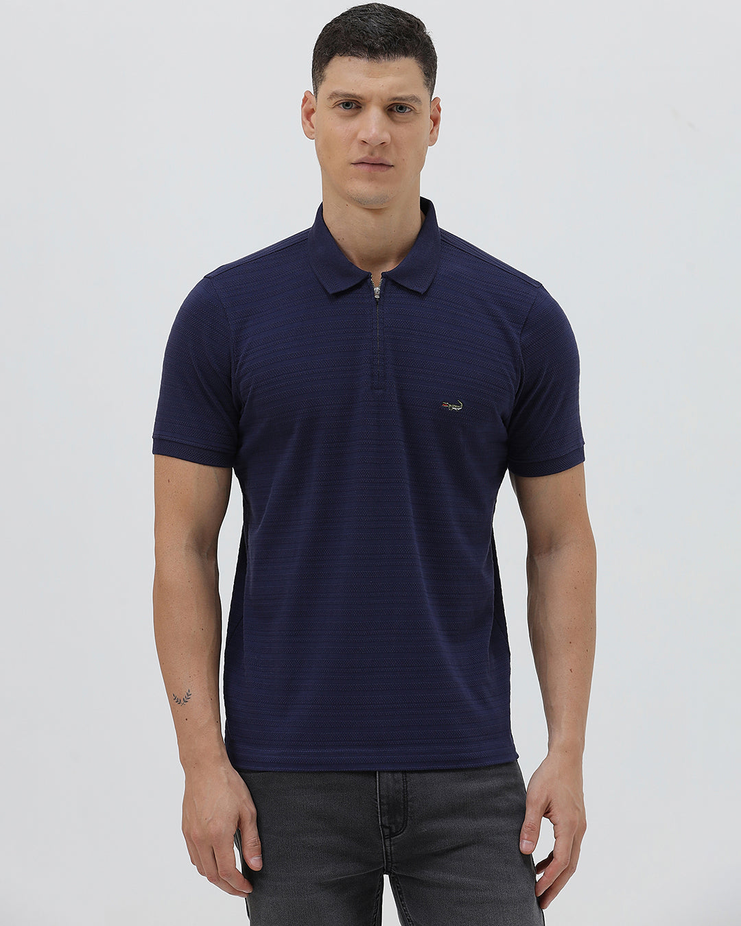 A man wearing a navy blue short-sleeve polo shirt with a small embroidered crocodile logo on the chest, paired with dark gray jeans, against a light gray studio background.
