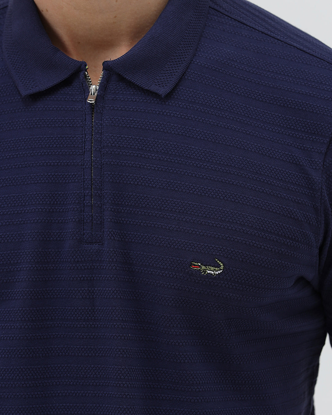 A man wearing a navy blue short-sleeve polo shirt with a small embroidered crocodile logo on the chest, paired with dark gray jeans, against a light gray studio background.