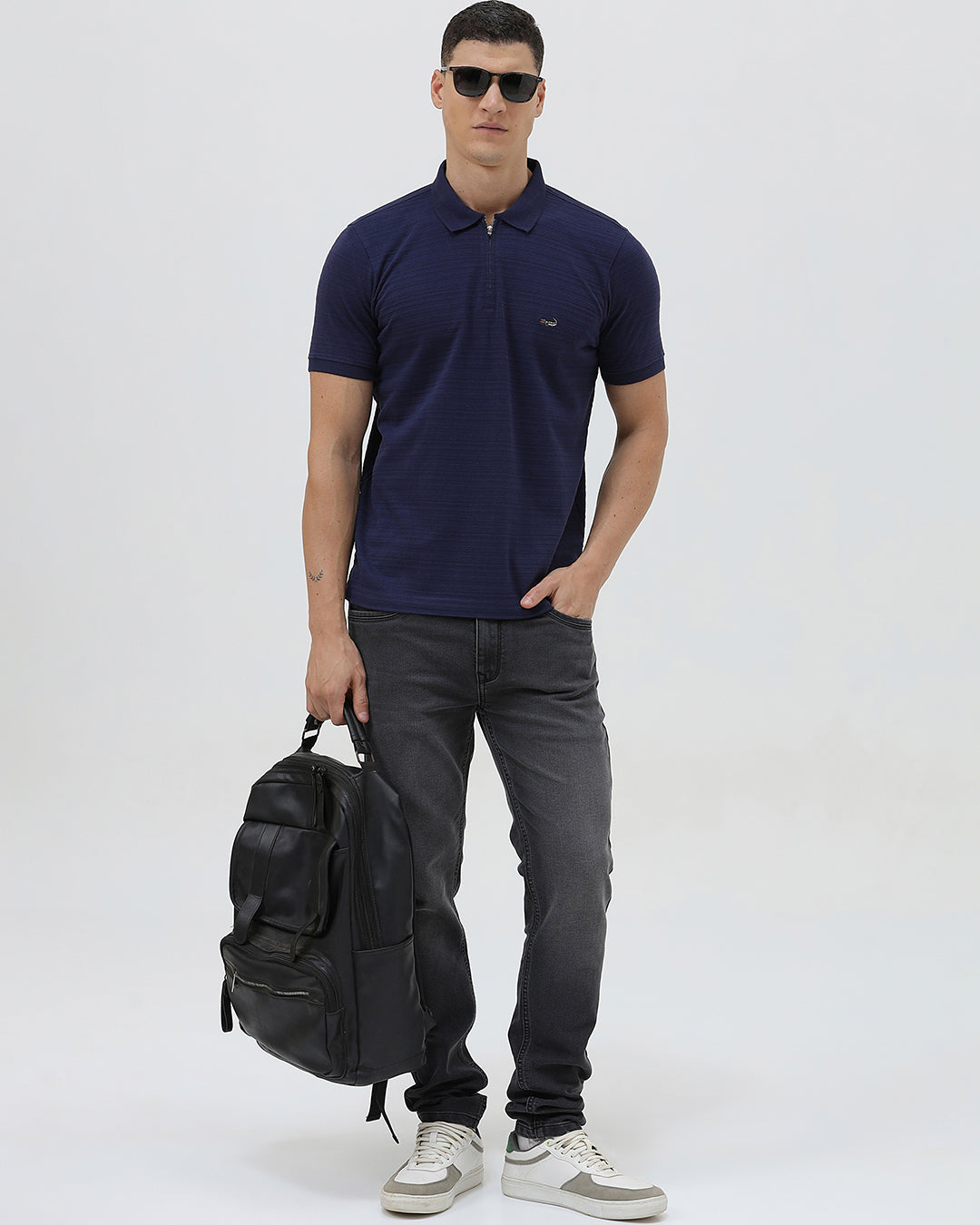A man wearing a navy blue short-sleeve polo shirt with a small embroidered crocodile logo on the chest, paired with dark gray jeans, against a light gray studio background.