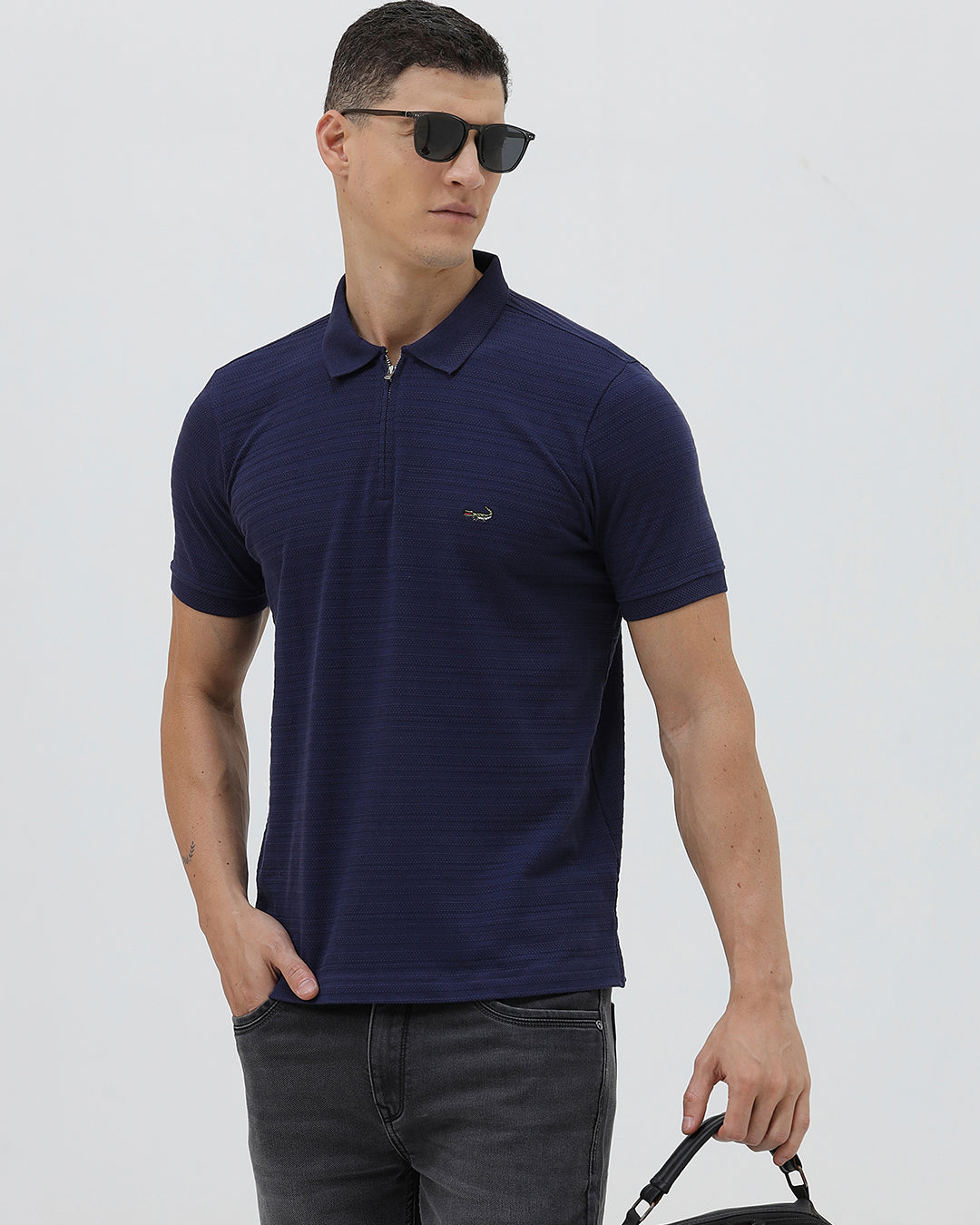A man wearing a navy blue short-sleeve polo shirt with a small embroidered crocodile logo on the chest, paired with dark gray jeans, against a light gray studio background.