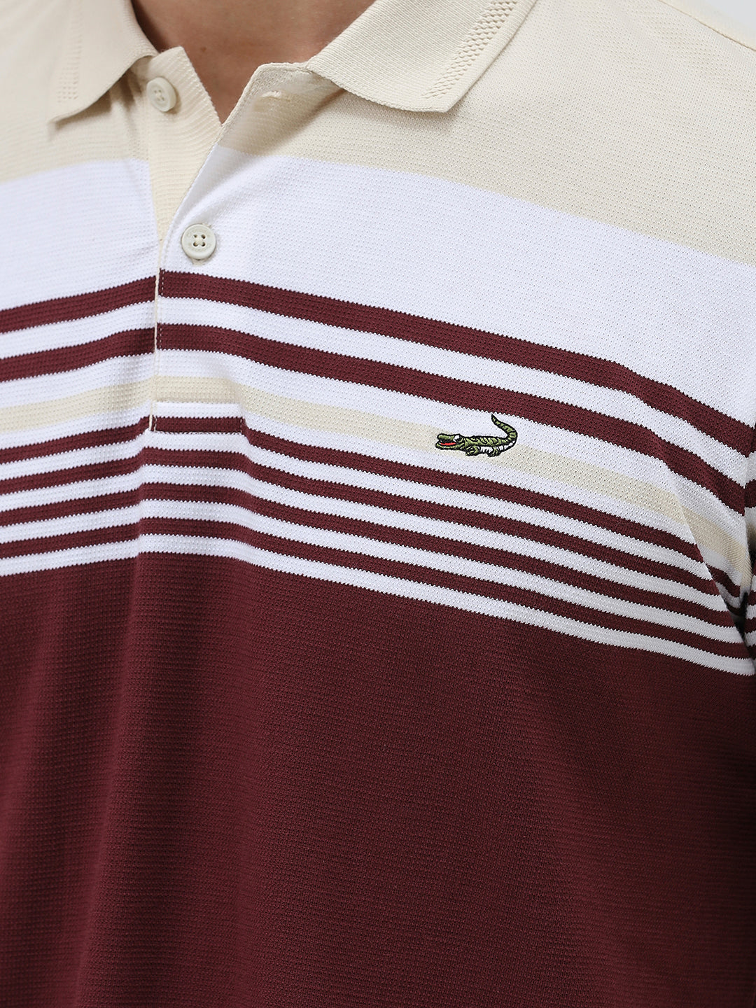 A man wearing a short-sleeve polo shirt with a colorblock design of cream, white, and burgundy, paired with dark blue jeans, against a plain light gray background.