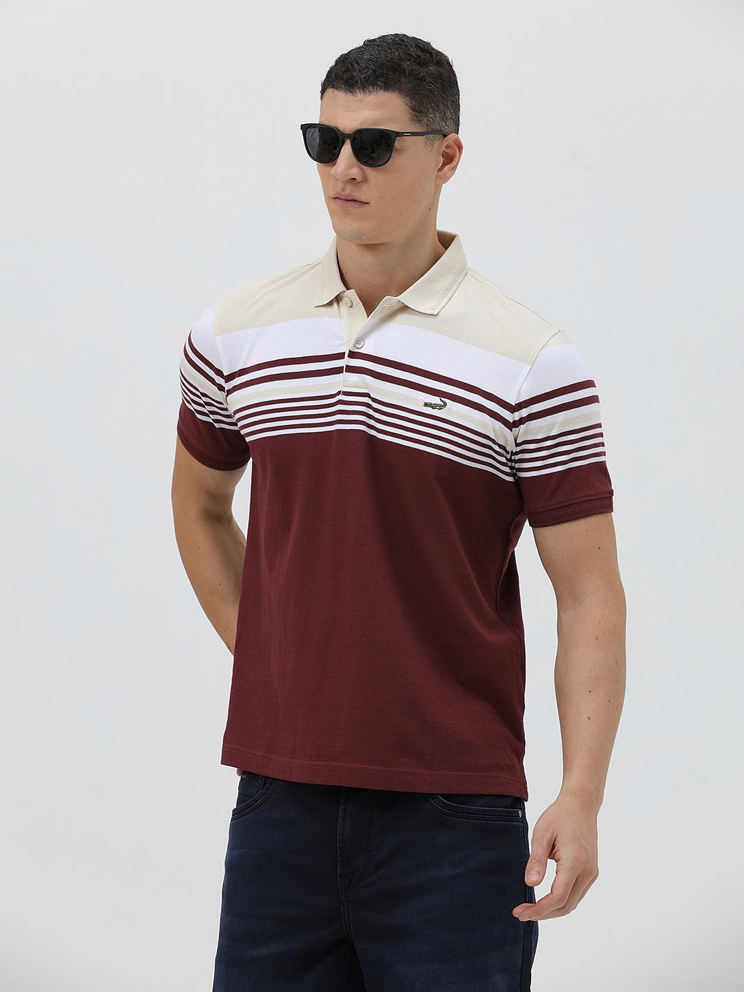 A man wearing a short-sleeve polo shirt with a colorblock design of cream, white, and burgundy, paired with dark blue jeans, against a plain light gray background.