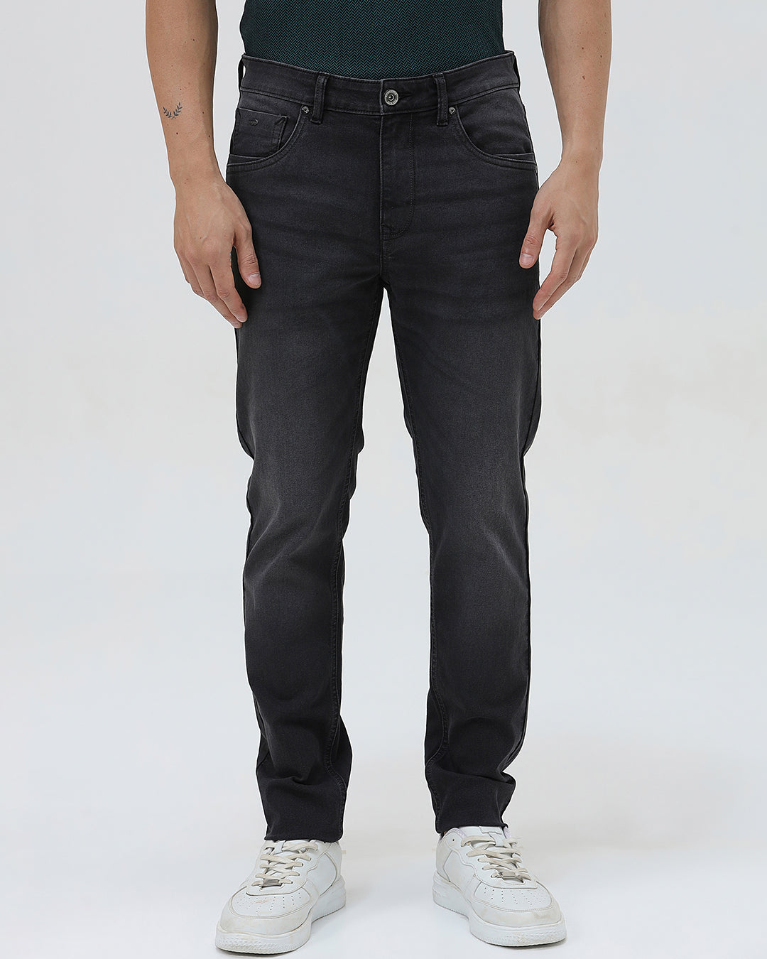 A person wearing slim-fit black denim jeans with a dark green top and white sneakers, shown from the waist down against a plain light gray studio background.