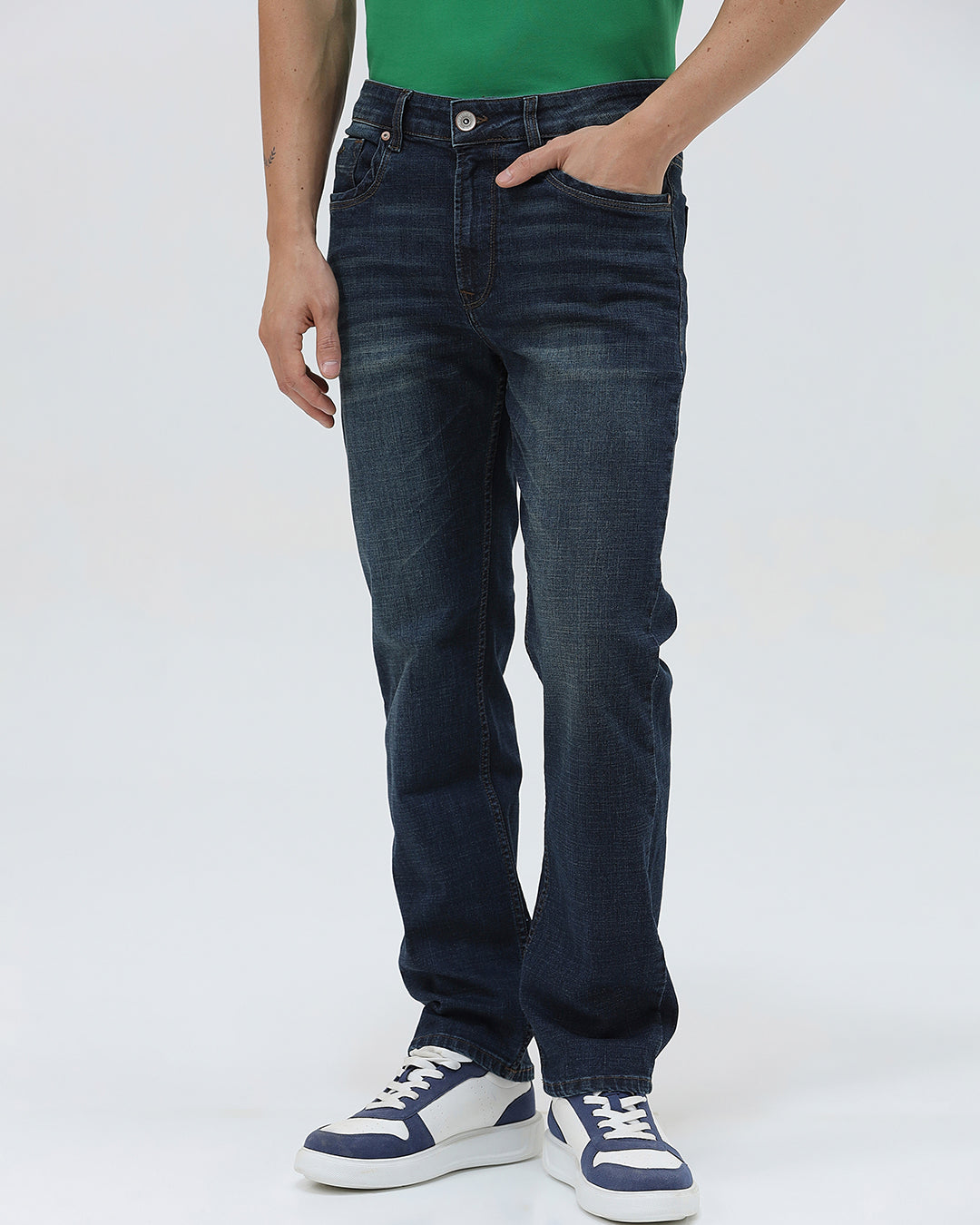 A person wearing dark blue slim-fit jeans, a green shirt, and navy and white sneakers, standing against a light gray studio background.