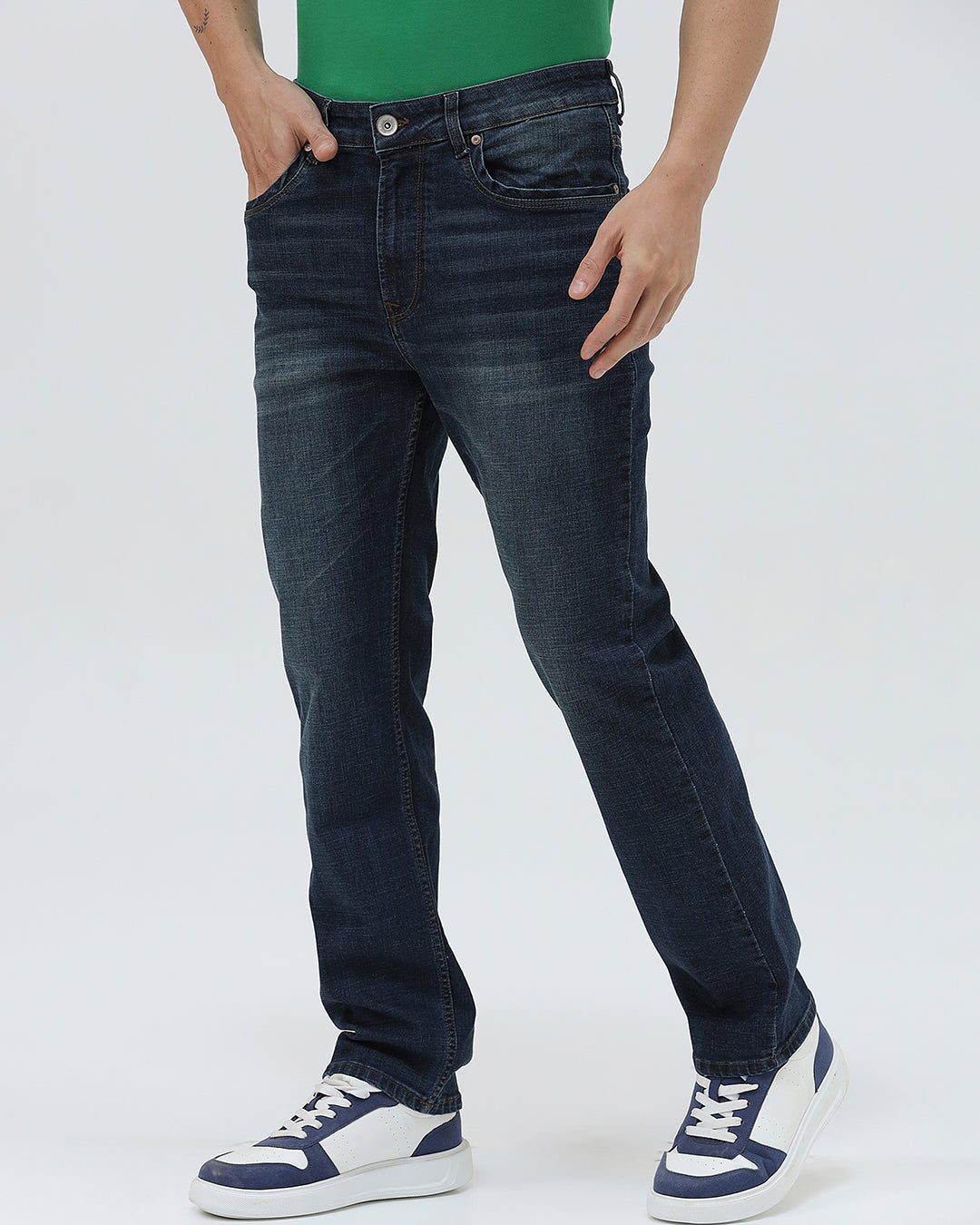 A person wearing dark blue slim-fit jeans, a green shirt, and navy and white sneakers, standing against a light gray studio background.