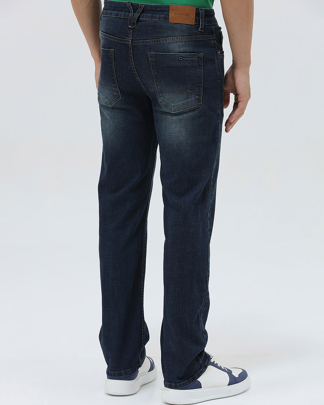 A person wearing dark blue slim-fit jeans, a green shirt, and navy and white sneakers, standing against a light gray studio background.