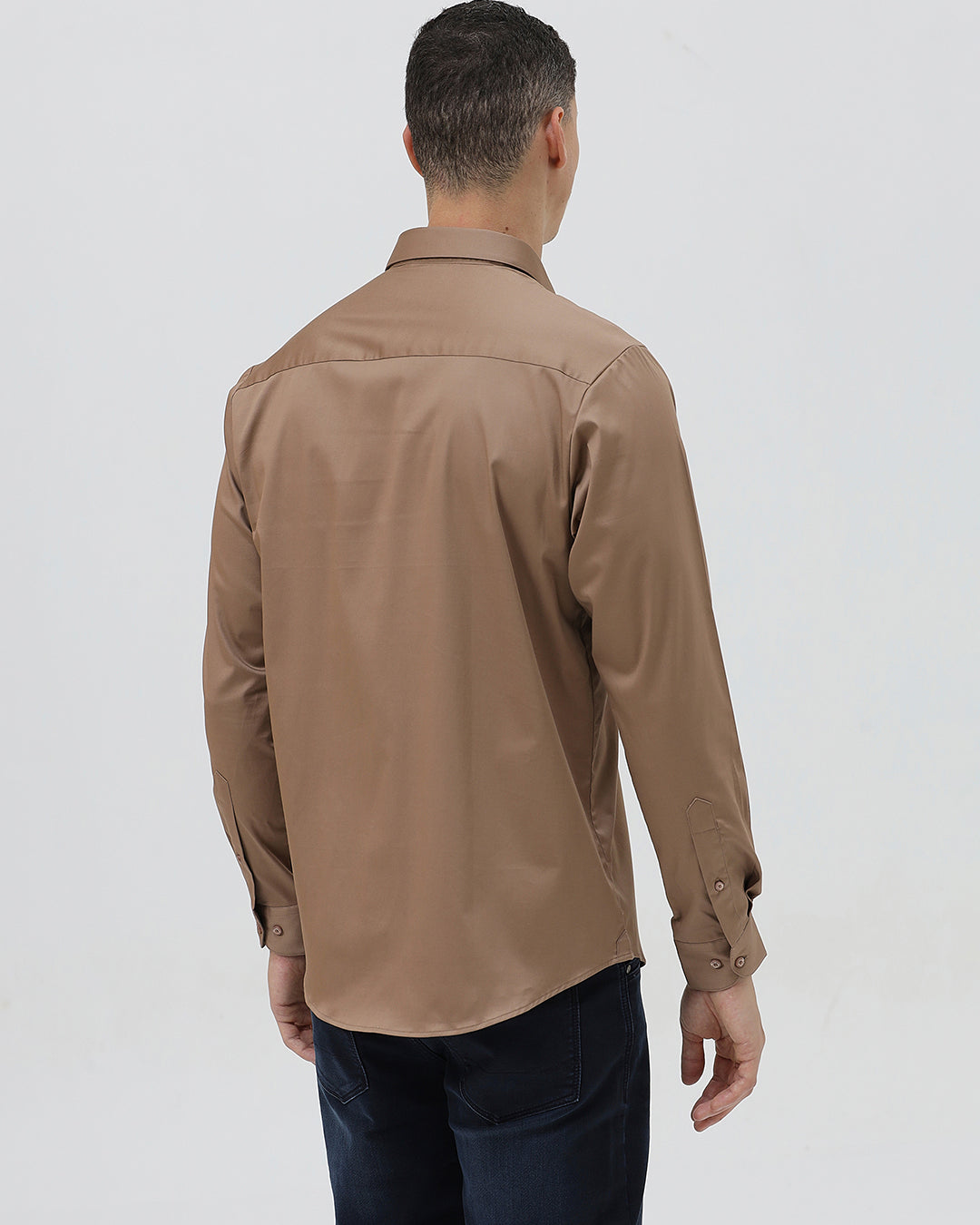 A man wearing a brown long-sleeve button-down shirt and dark blue jeans, standing against a light gray studio background.