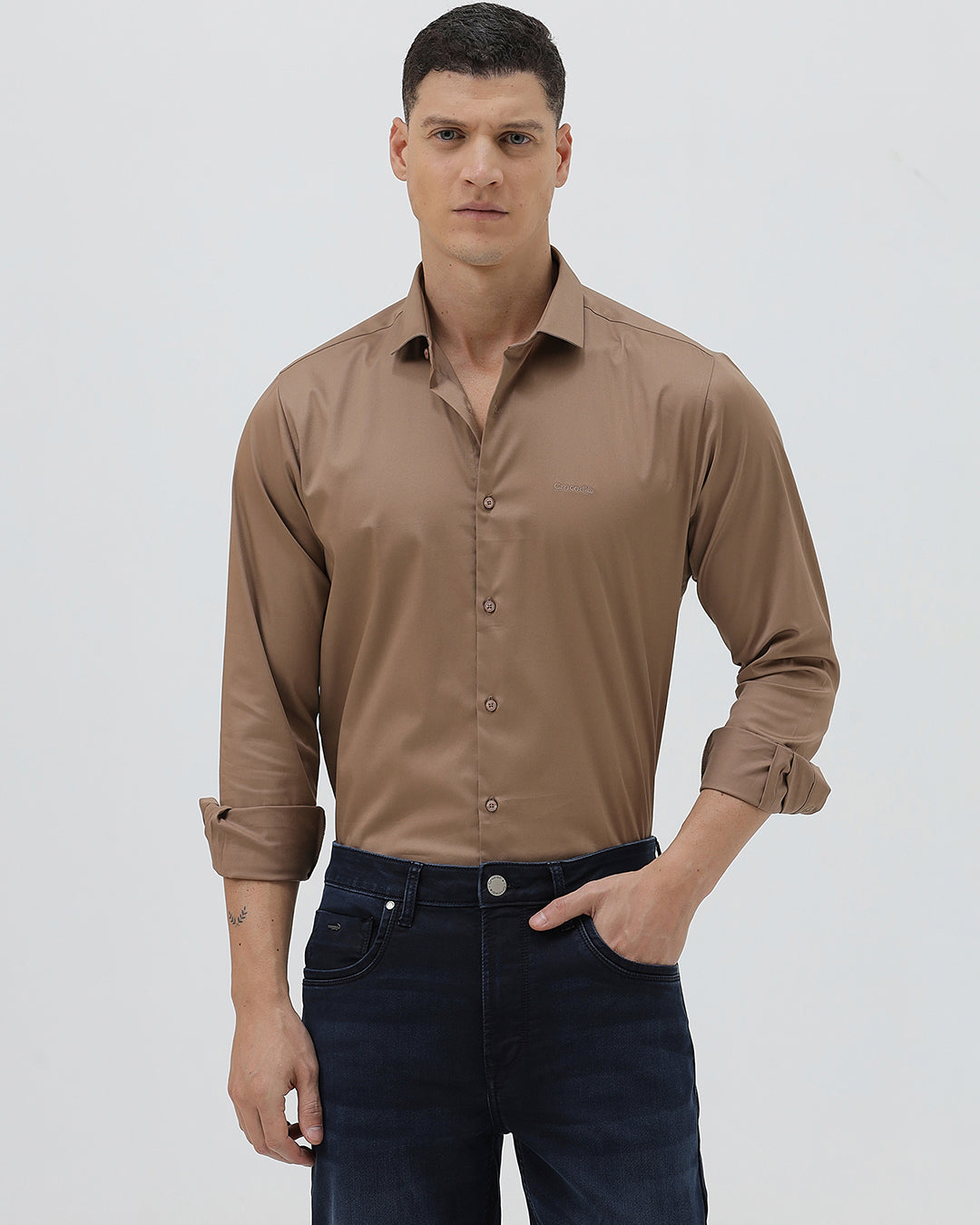 A man wearing a brown long-sleeve button-down shirt and dark blue jeans, standing against a light gray studio background.