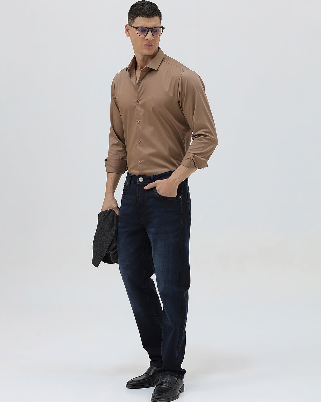 A man wearing a brown long-sleeve button-down shirt and dark blue jeans, standing against a light gray studio background.