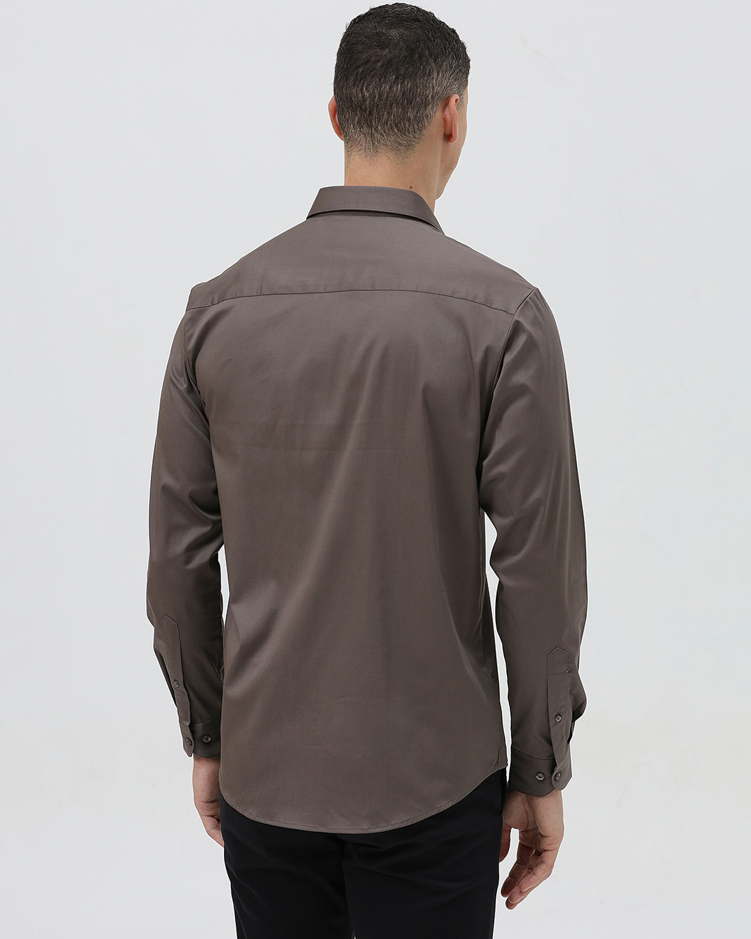 A man wearing a long-sleeve button-down dress shirt in a dark taupe-brown color, paired with black trousers, photographed against a light gray studio background.
