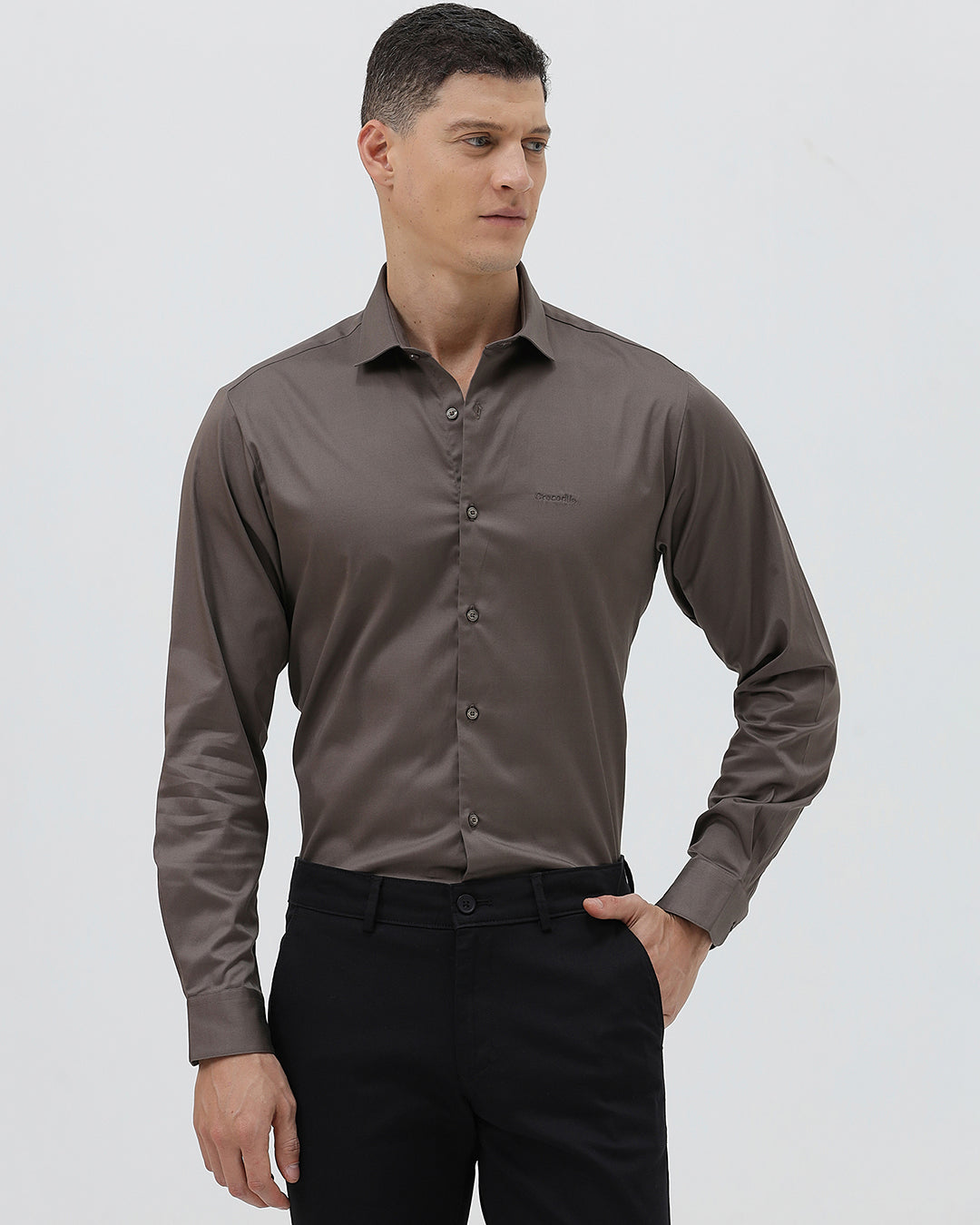 A man wearing a long-sleeve button-down dress shirt in a dark taupe-brown color, paired with black trousers, photographed against a light gray studio background.
