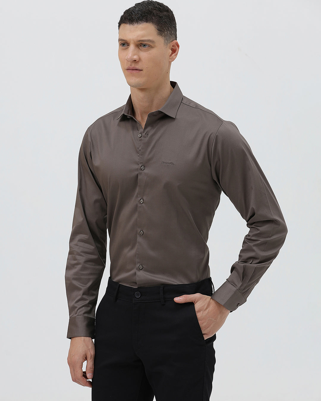 A man wearing a long-sleeve button-down dress shirt in a dark taupe-brown color, paired with black trousers, photographed against a light gray studio background.