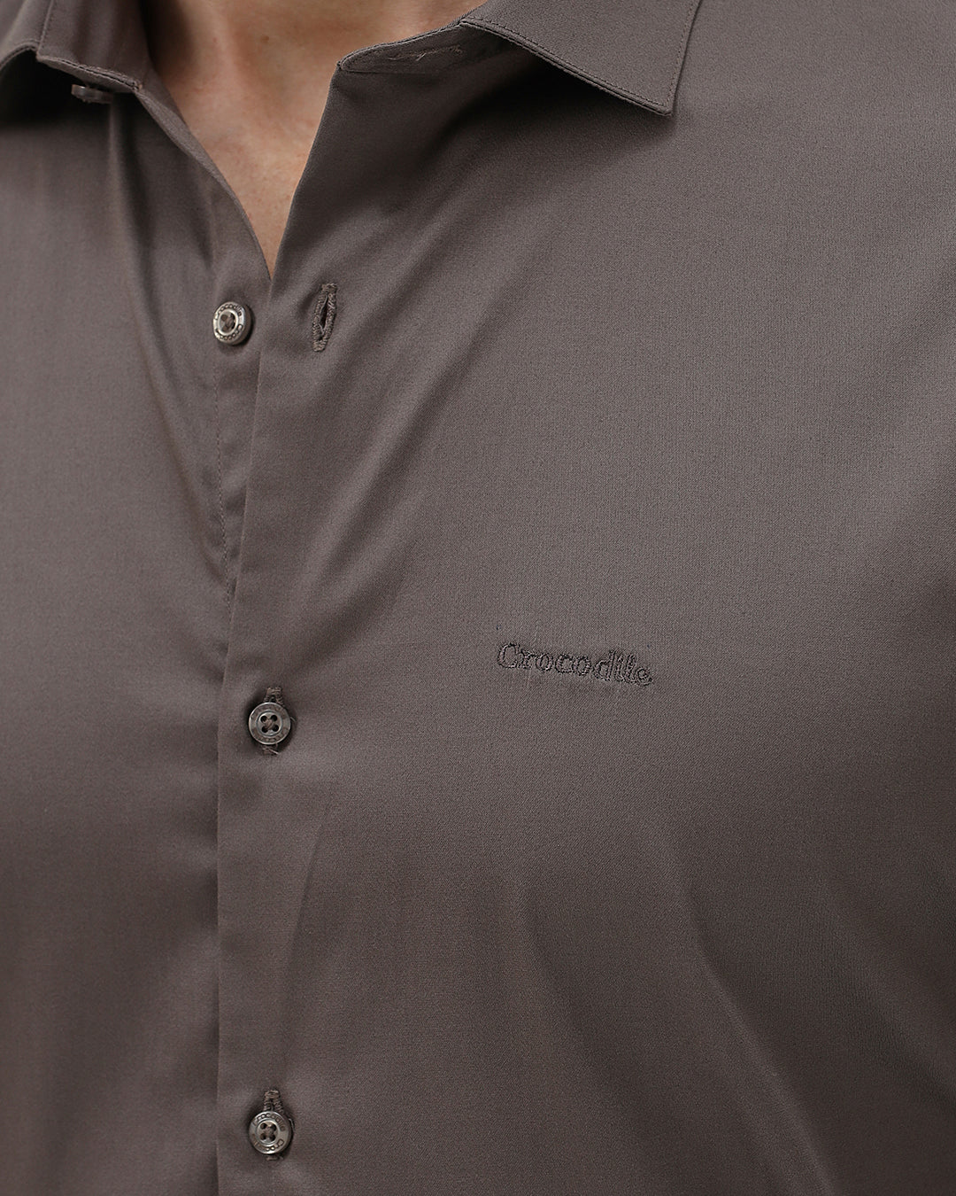 A man wearing a long-sleeve button-down dress shirt in a dark taupe-brown color, paired with black trousers, photographed against a light gray studio background.