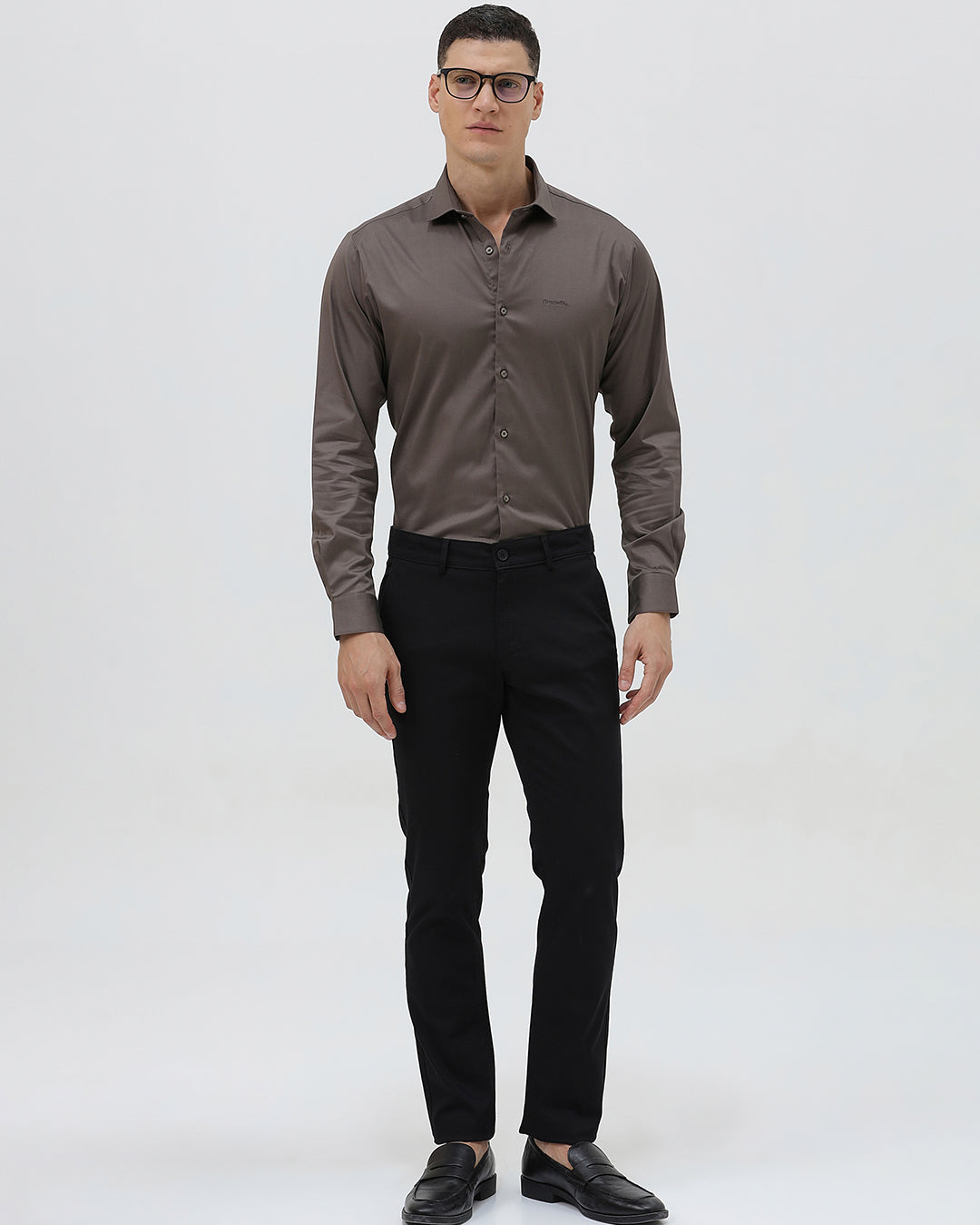 A man wearing a long-sleeve button-down dress shirt in a dark taupe-brown color, paired with black trousers, photographed against a light gray studio background.