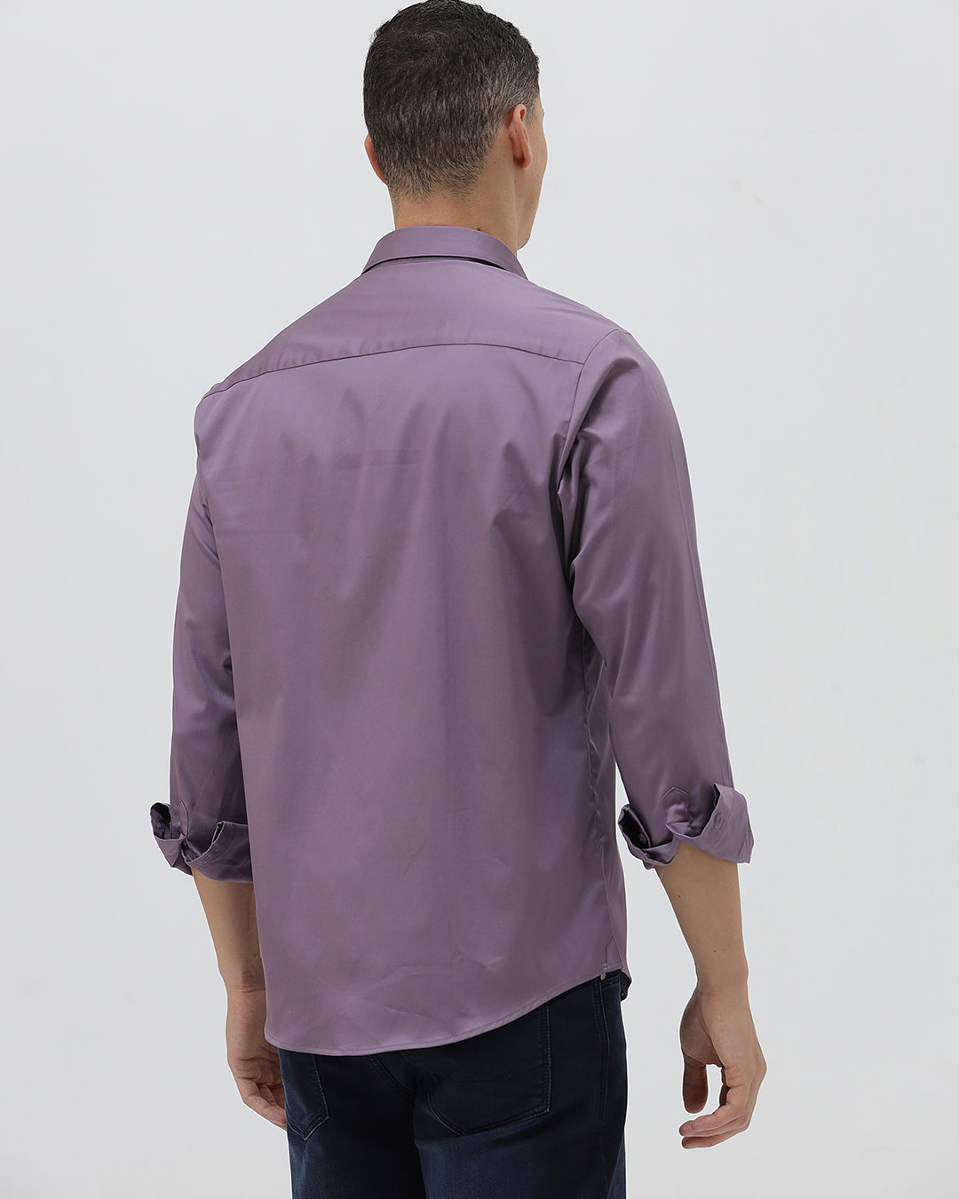 A man wearing a long-sleeve button-down shirt in a muted purple shade, paired with dark blue jeans, standing against a light gray studio background.
