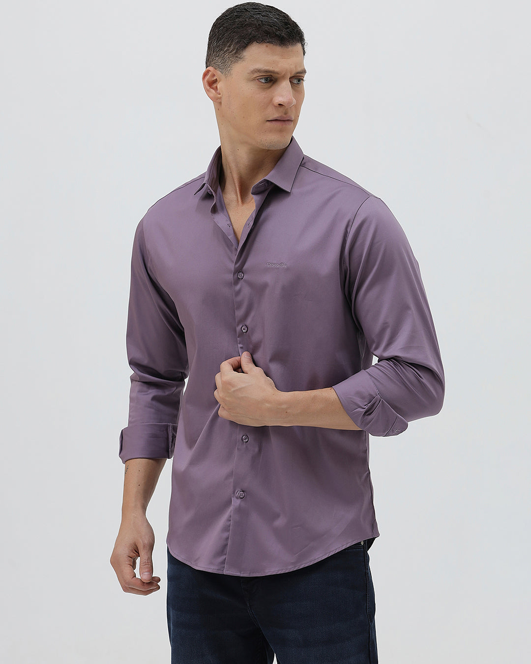 A man wearing a long-sleeve button-down shirt in a muted purple shade, paired with dark blue jeans, standing against a light gray studio background.