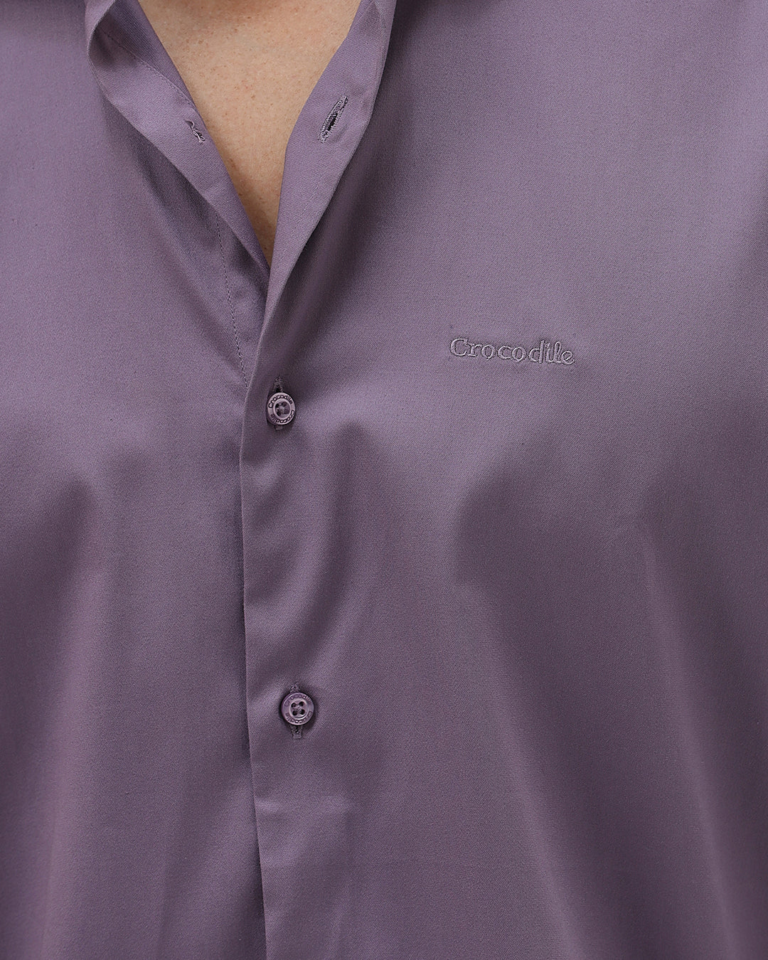 A man wearing a long-sleeve button-down shirt in a muted purple shade, paired with dark blue jeans, standing against a light gray studio background.