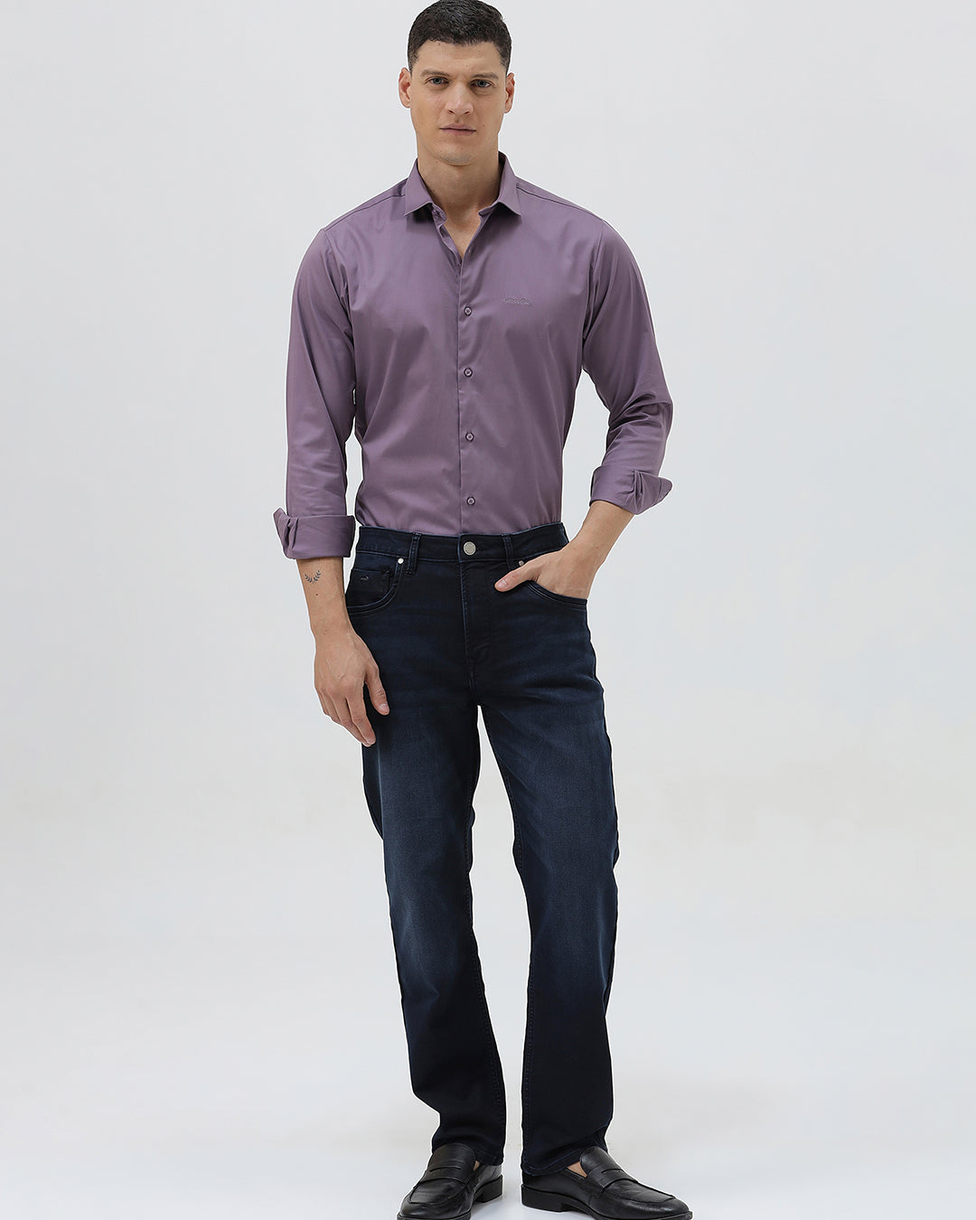 A man wearing a long-sleeve button-down shirt in a muted purple shade, paired with dark blue jeans, standing against a light gray studio background.