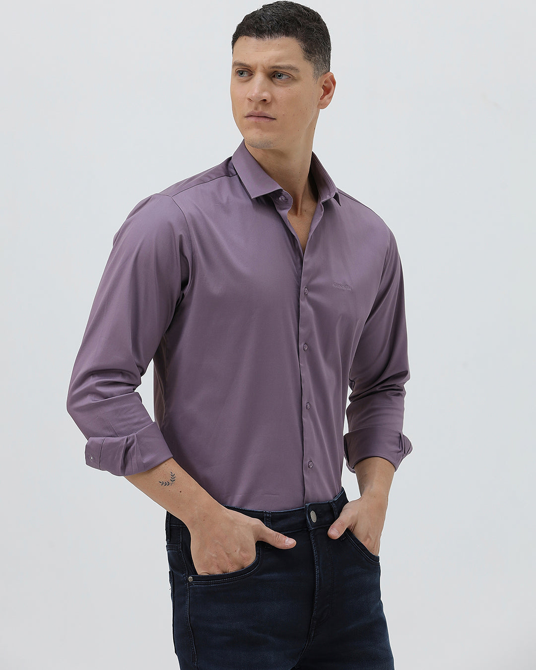 A man wearing a long-sleeve button-down shirt in a muted purple shade, paired with dark blue jeans, standing against a light gray studio background.