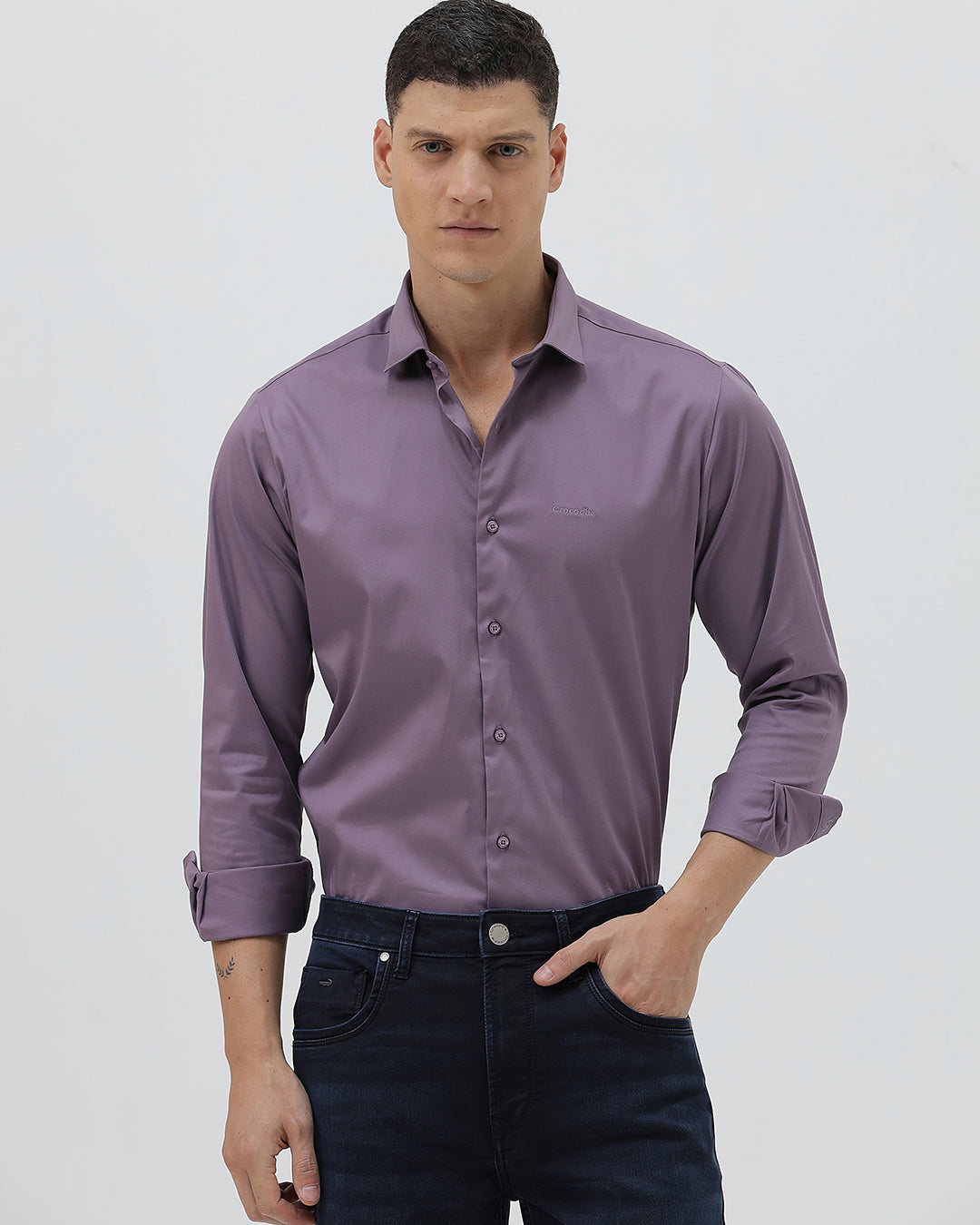 A man wearing a long-sleeve button-down shirt in a muted purple shade, paired with dark blue jeans, standing against a light gray studio background.