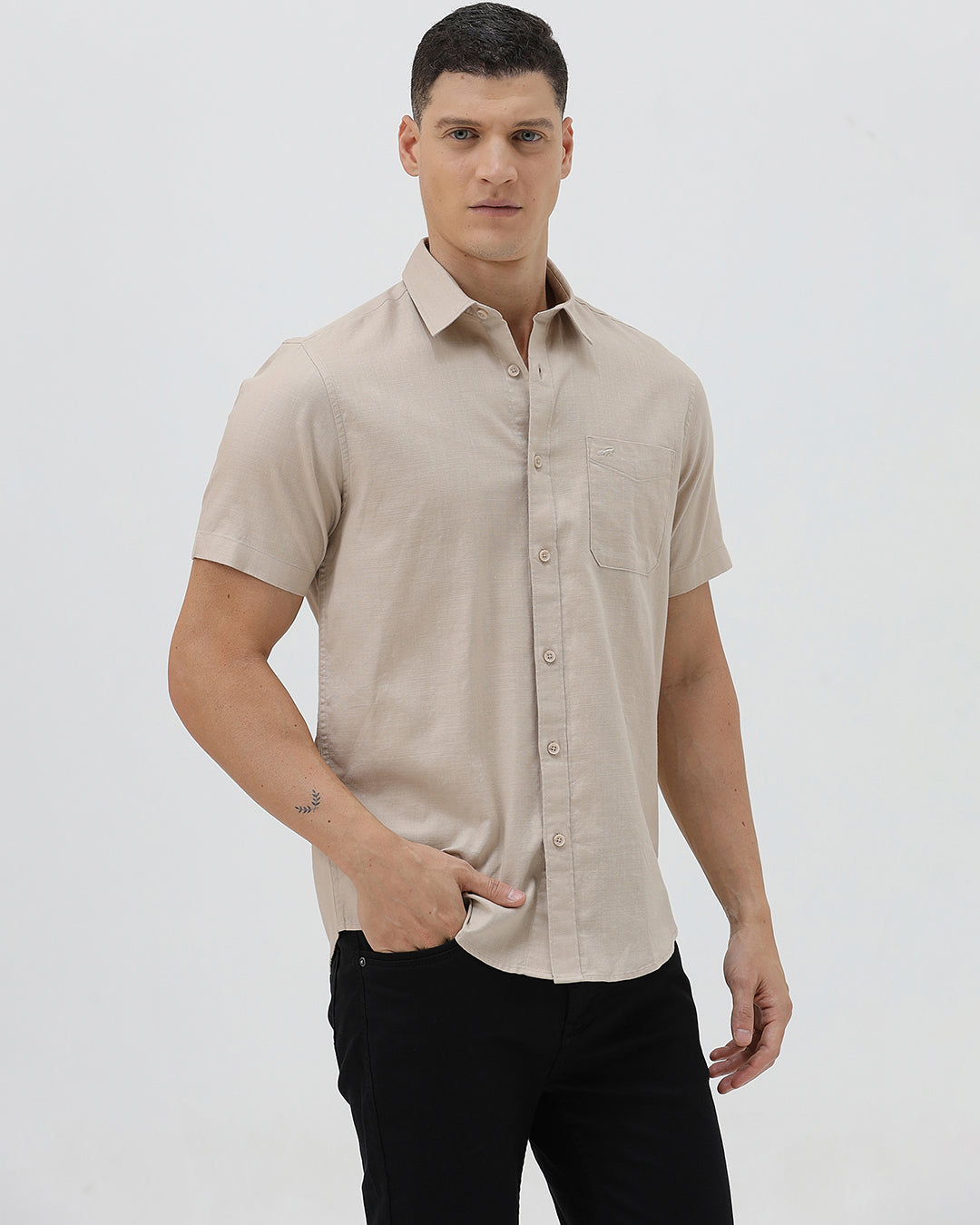 A man wearing a dusty rose long-sleeve button-down shirt and light beige trousers, standing against a light gray studio background.