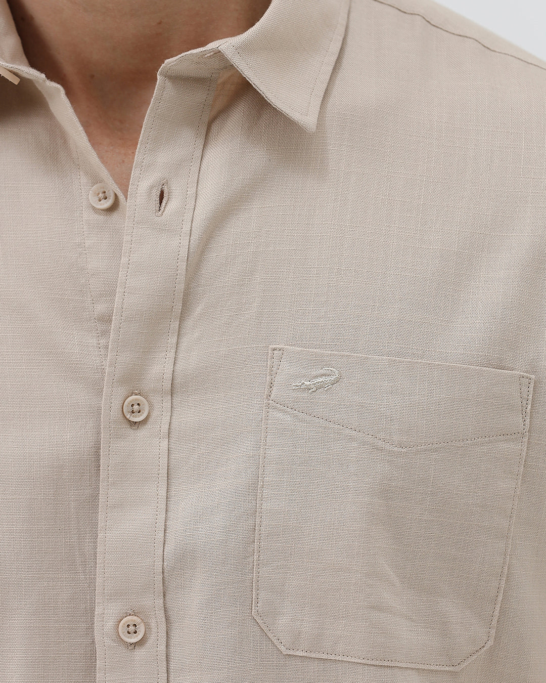 A man wearing a dusty rose long-sleeve button-down shirt and light beige trousers, standing against a light gray studio background.