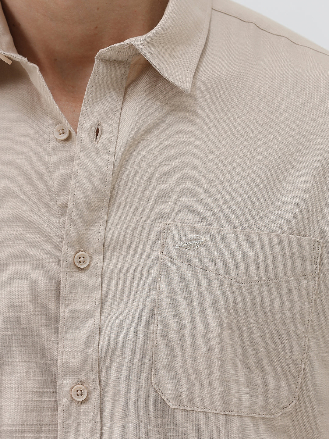 A man wearing a dusty rose long-sleeve button-down shirt and light beige trousers, standing against a light gray studio background.