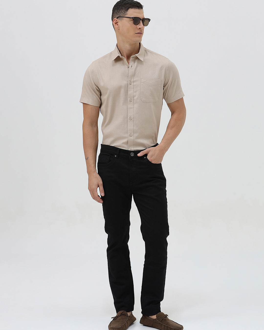 A man wearing a dusty rose long-sleeve button-down shirt and light beige trousers, standing against a light gray studio background.
