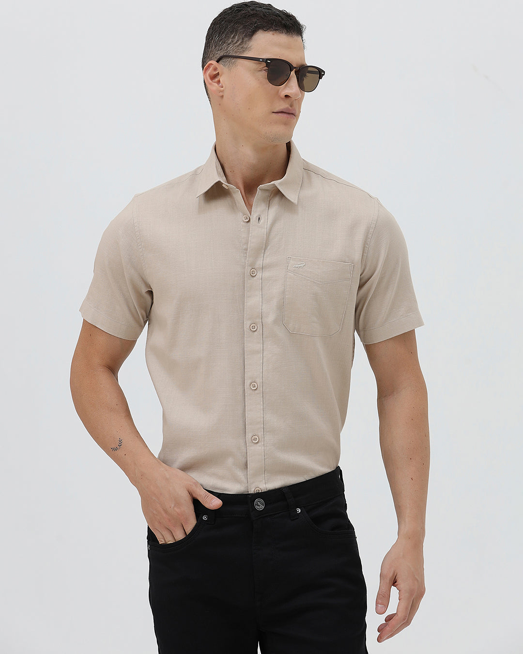 A man wearing a dusty rose long-sleeve button-down shirt and light beige trousers, standing against a light gray studio background.