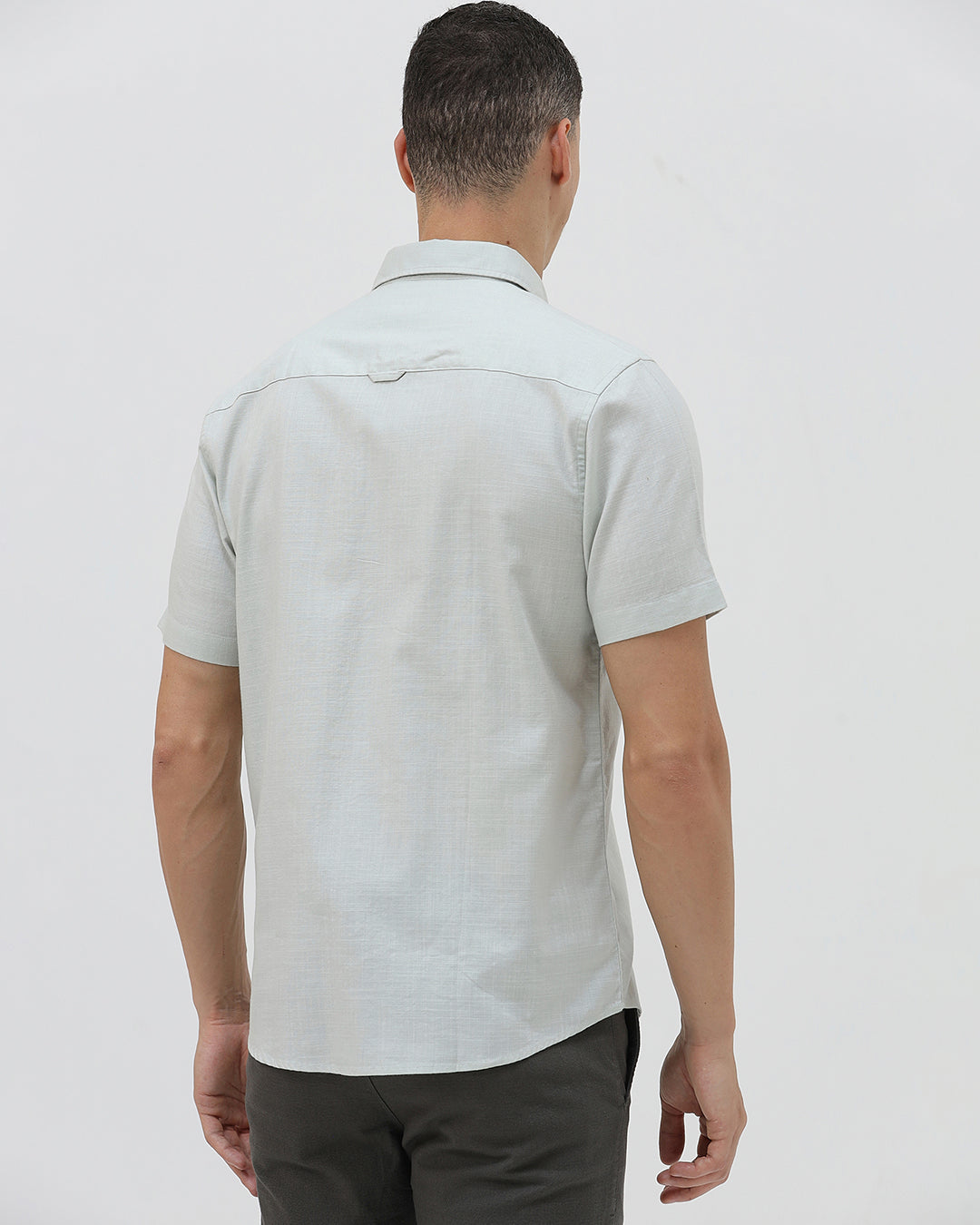 A man wearing a light sage green short-sleeve button-down shirt and dark gray trousers, standing against a plain light gray studio background.